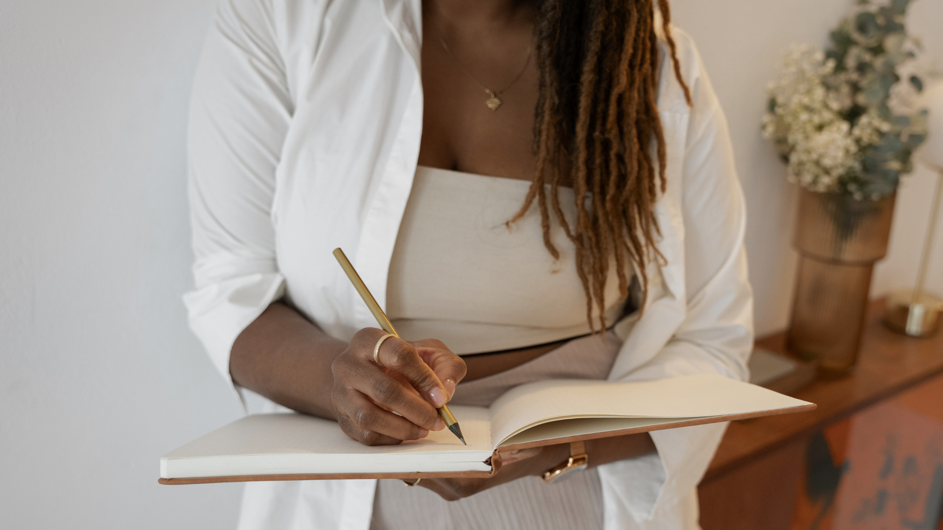 Person writing in a notebook with a pen, wearing a white outfit, next to a vase of flowers.