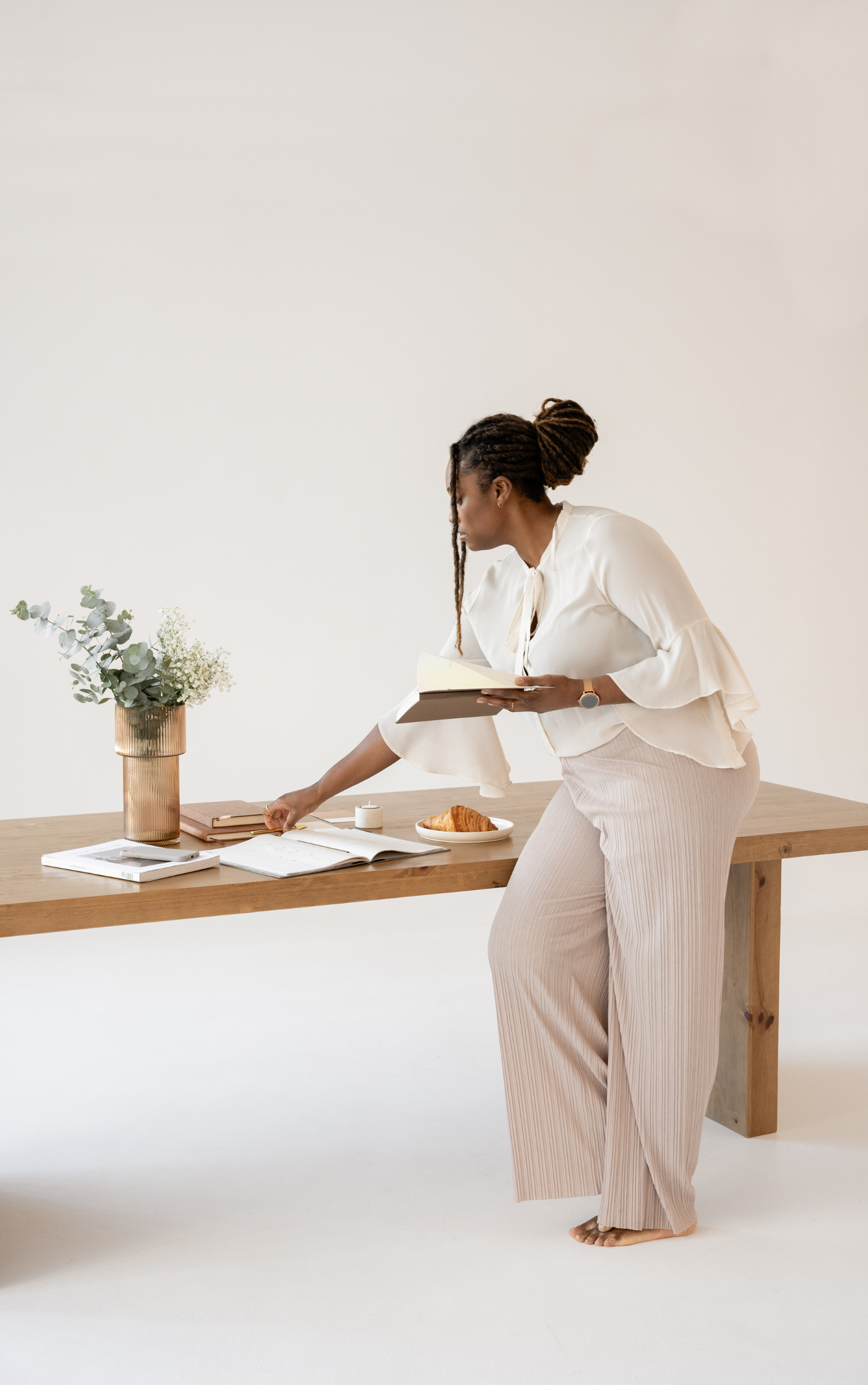 Person organizing a wooden table with books, a vase of flowers, and a croissant.