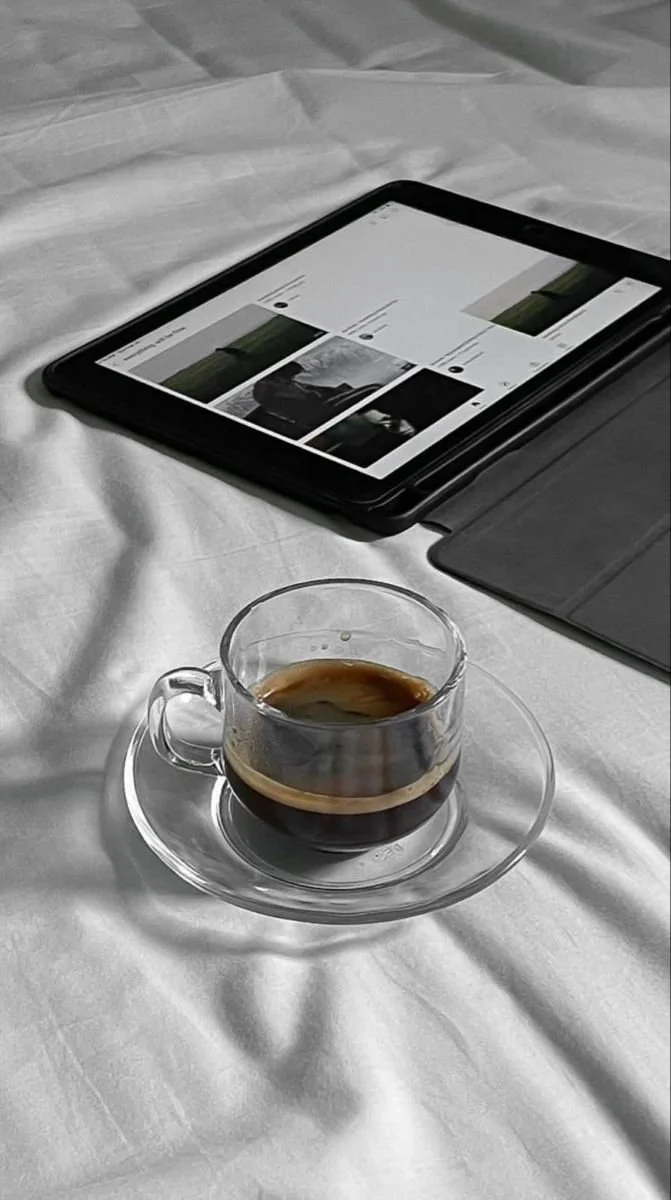 A glass cup of coffee on a saucer with a dark-colored tablet or e-reader and a paper napkin on a white tablecloth.
