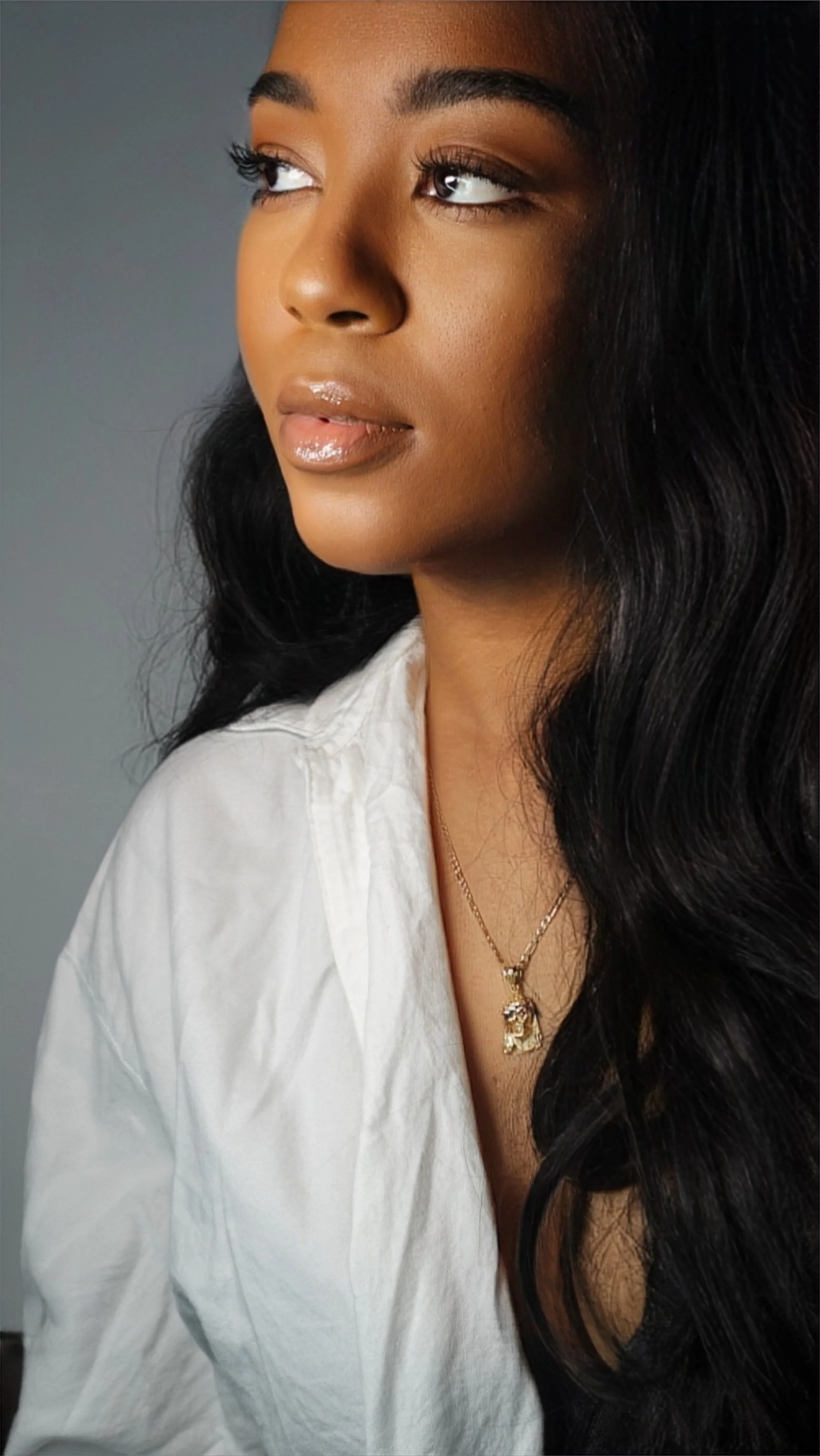 Close-up portrait of a woman with long black curly hair, wearing a white shirt and a gold necklace with a figure pendant, looking to the side against a grey background.