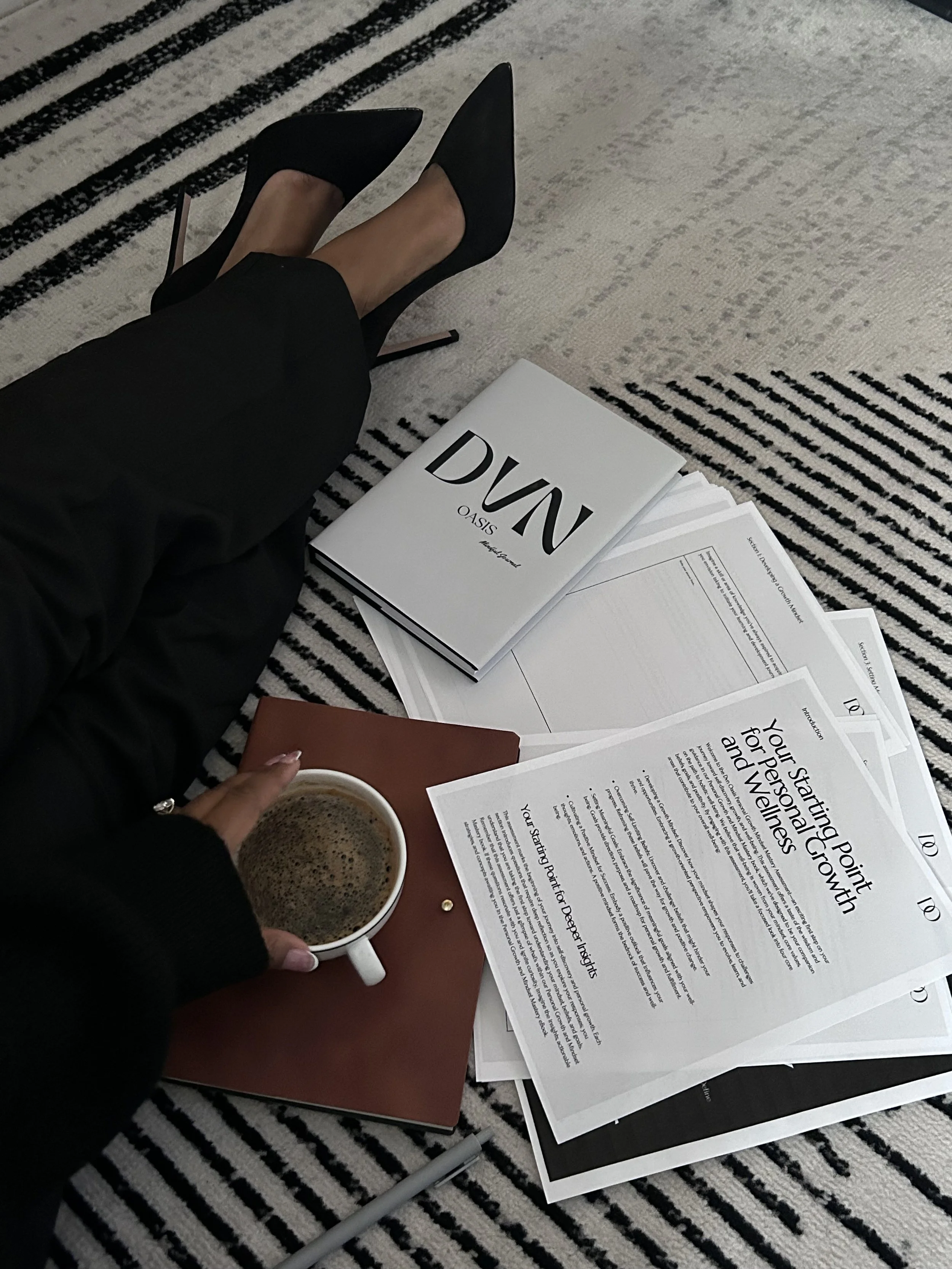 A person with black high heels and black pants sitting on a striped rug, holding a cup of coffee, with papers, notebooks, and magazines spread out on the floor.