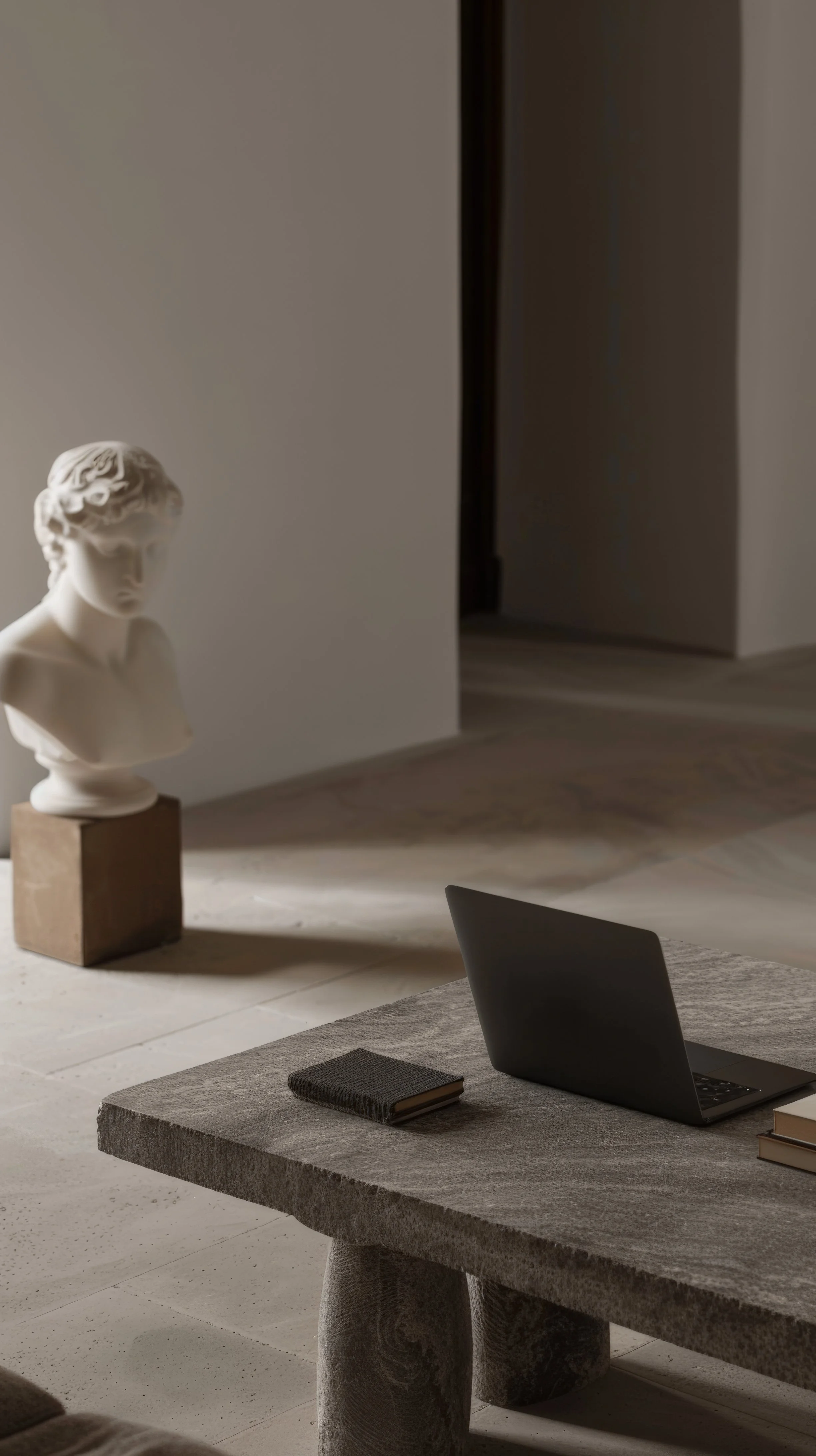 A modern interior with a gray stone table, a laptop, a black notebook, and a closed book. In the background, a classical marble bust sculpture of a woman's head is displayed on a wooden block. The room has minimalist decor with a neutral color palette.