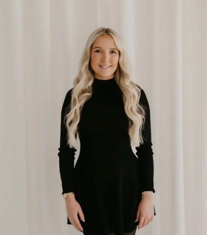A young woman with long blonde hair wearing a black long-sleeve dress smiling at the camera, standing in front of a light-colored background.