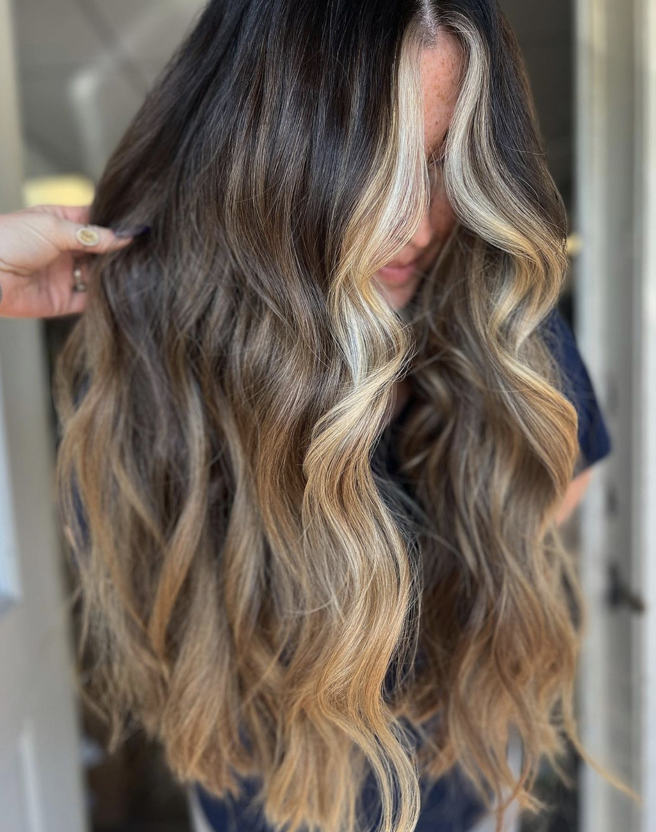 Close-up of a woman with long, wavy hair featuring dark roots and blonde highlights, styled in loose waves.
