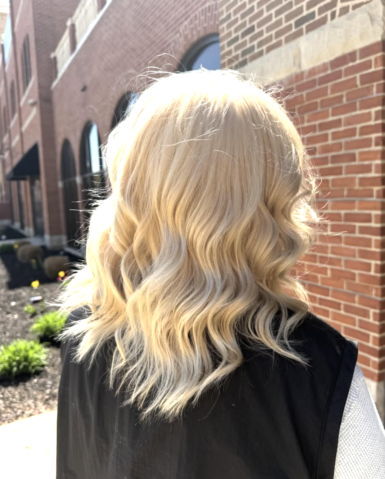 Back view of a woman with long, wavy blonde hair standing outside near a brick building with arched windows and a small garden bed.