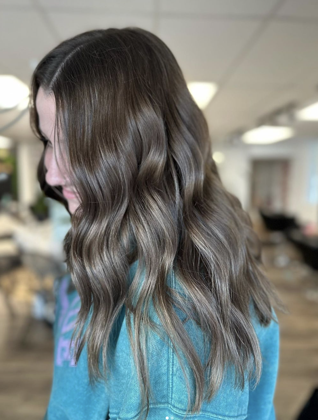 Woman with long, wavy, brown hair in a salon or indoor setting, wearing a blue shirt.