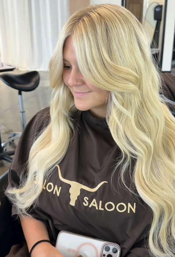 Woman with long blonde wavy hair sitting in salon chair, wearing a salon cape with longhorn logo.