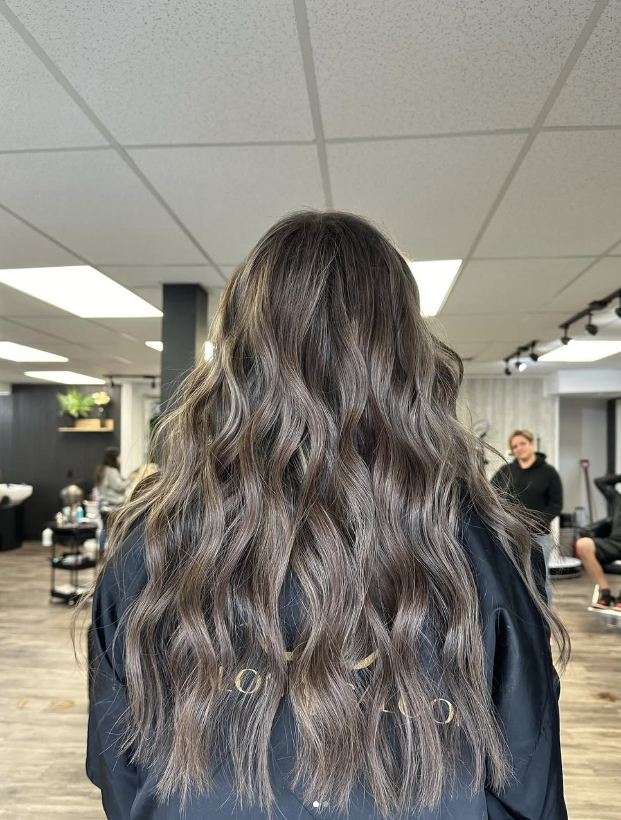 Back of a person's head showcasing long, wavy, ombre hair transitioning from dark brown to ash blonde in a salon setting.