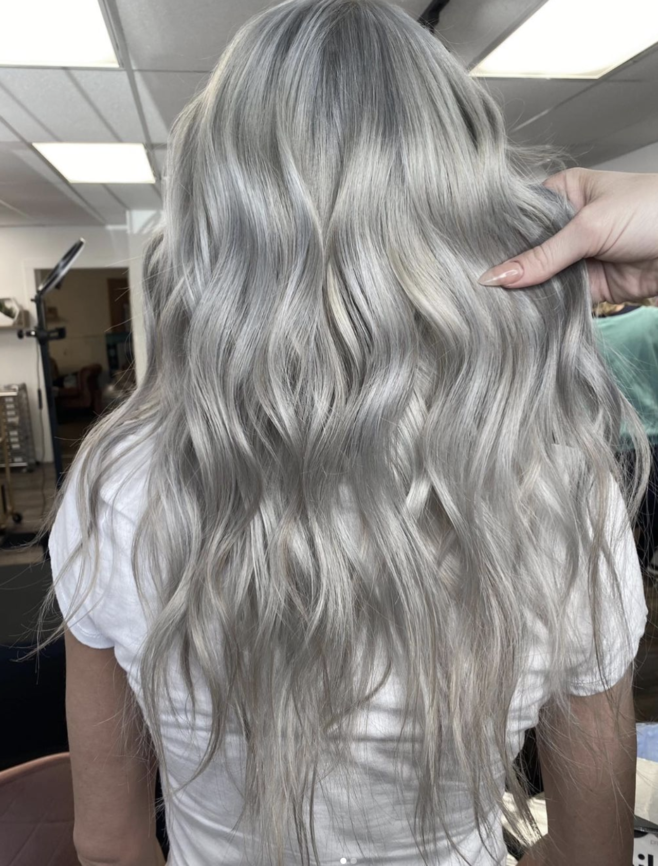 A person with long, wavy, platinum blonde hair, partially held by a hand at the side of the head.