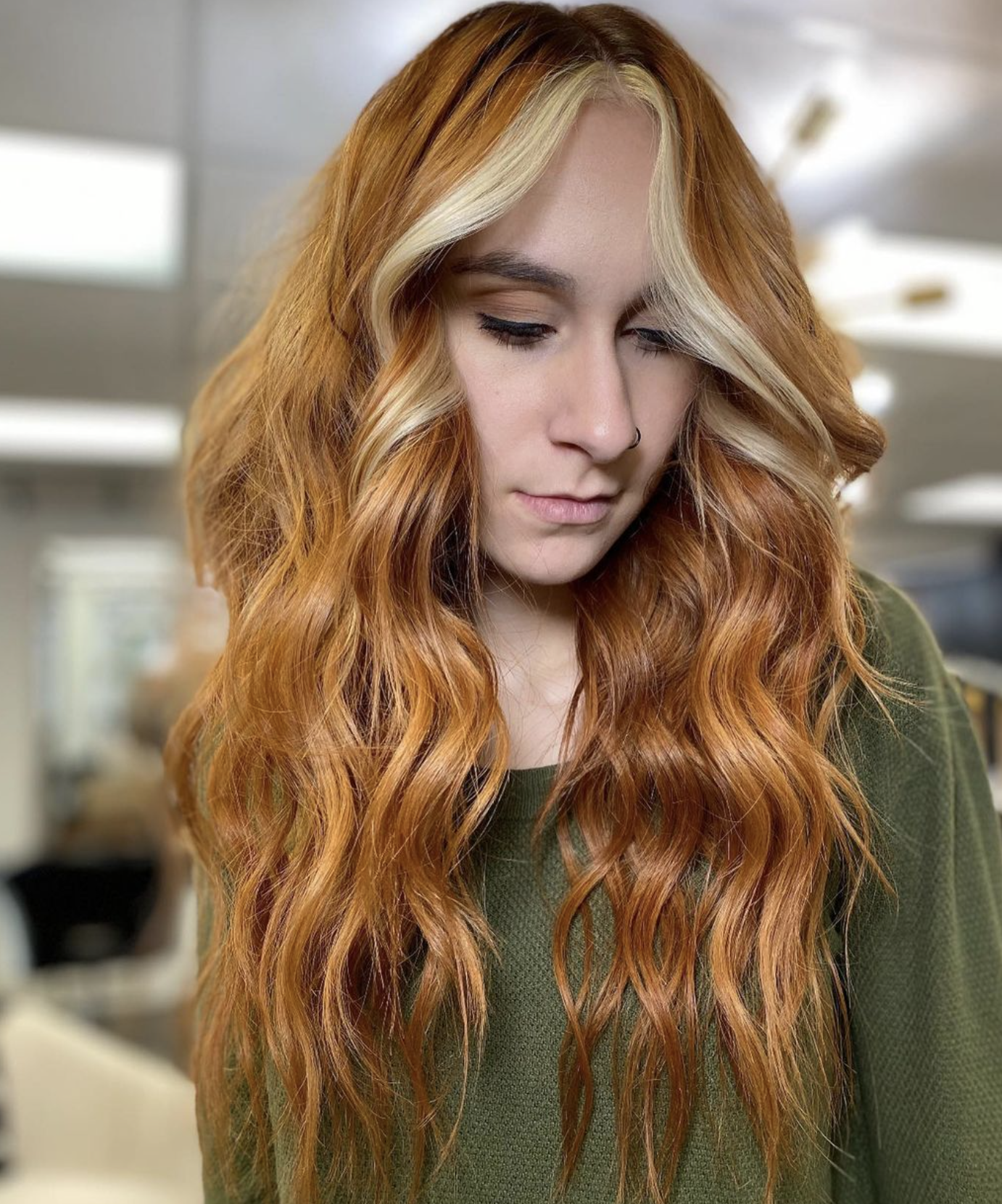 A young woman with long, wavy red hair and blonde highlights, wearing a nose ring and a dark green sweater, looking down in a brightly lit indoor space.