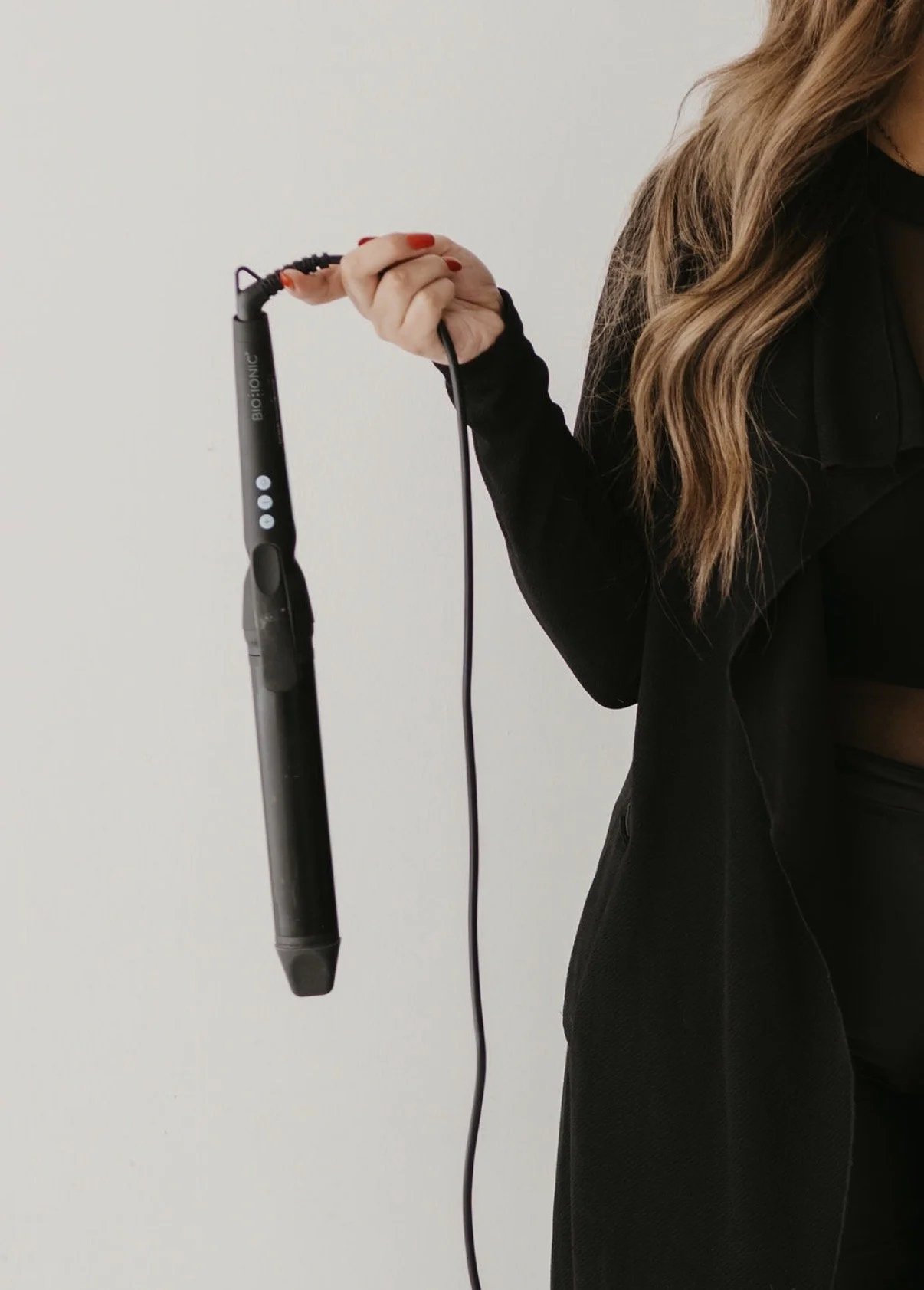 A woman with long wavy hair wearing a black blazer and a sheer black top holding a black Biolonic hair styling tool with a cord against a plain white background.