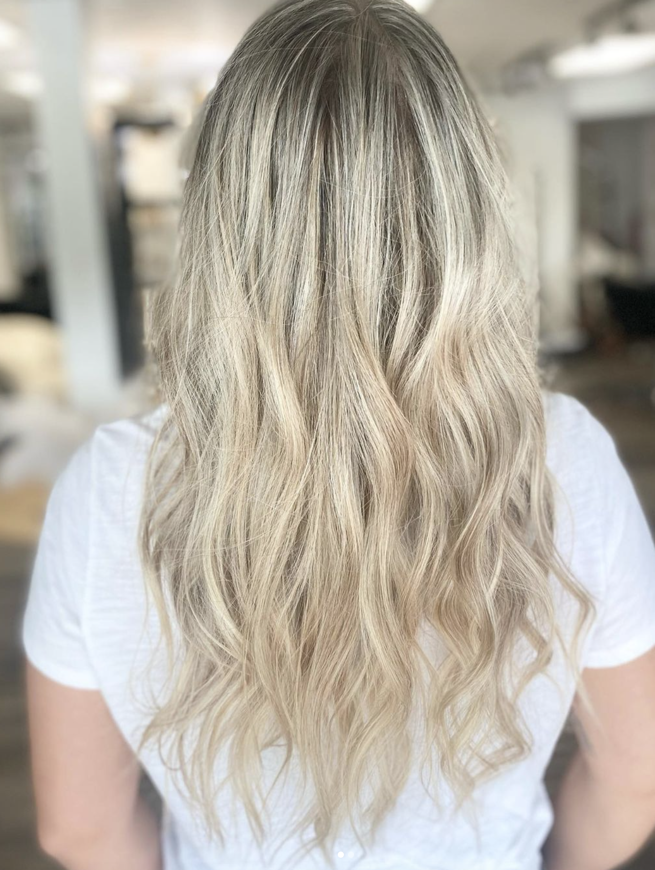 Back view of a woman with long, wavy blonde hair wearing a white shirt in a bright interior space.