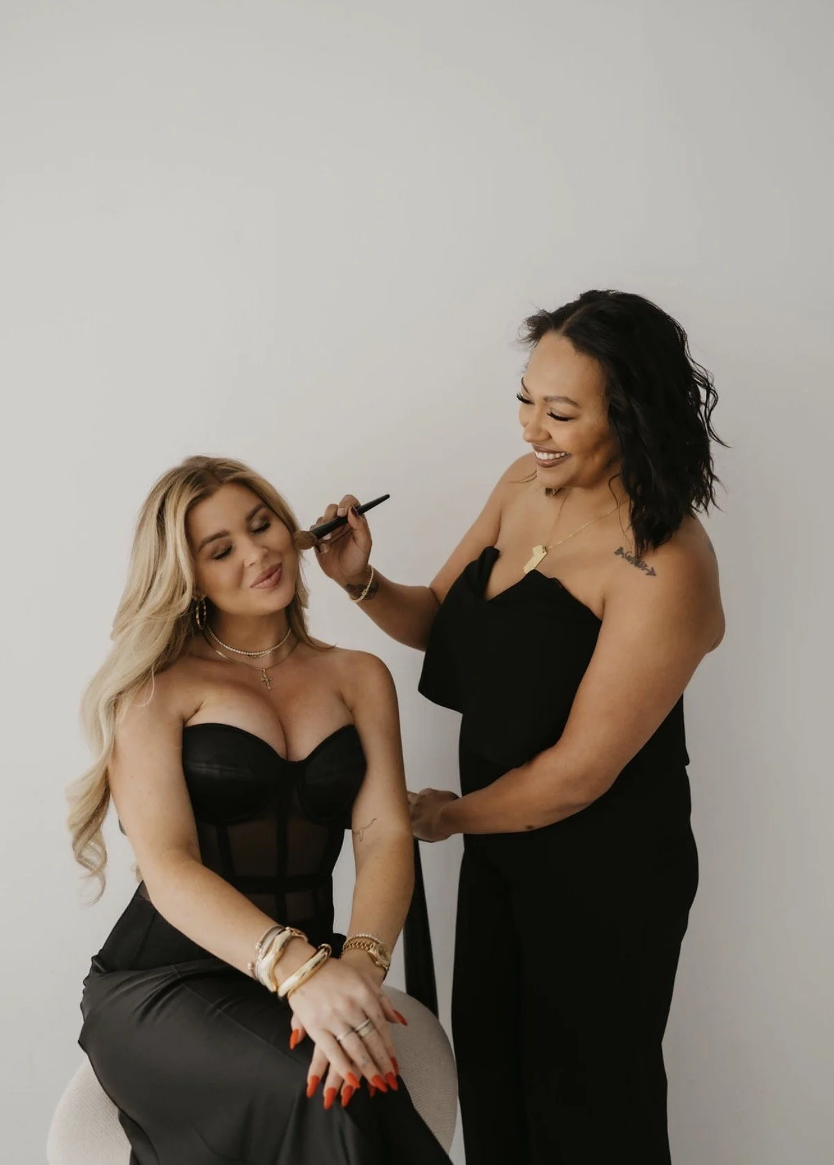 A woman with long blonde hair, wearing a black strapless top and a leather skirt, sitting with her eyes closed while a makeup artist applies makeup to her face. The makeup artist has dark hair, is smiling, and is wearing a black strapless dress. They are standing against a plain white background.