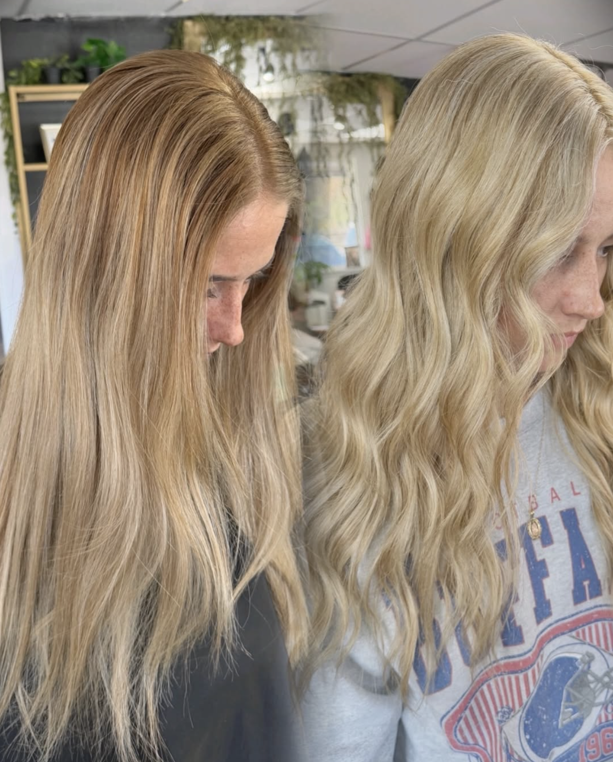 Side view of two women with long, wavy blonde hair, one with slightly darker blonde hair on the left and the other with lighter blonde hair on the right, in an indoor setting with plants and shelves in the background.