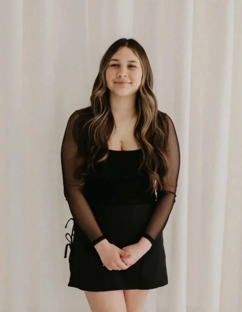 A young woman with long, wavy brown hair, wearing a black dress with sheer sleeves, standing in front of white curtains and smiling softly.