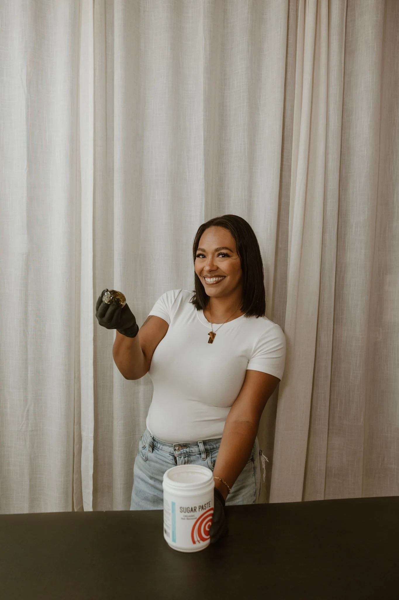 A woman with shoulder-length black curly hair, smiling, wearing a beige blazer over a black top, and a gold necklace with a number one pendant, standing against a neutral background.