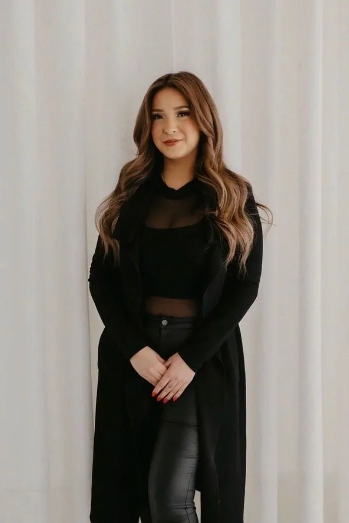 A woman with long wavy brown hair wearing a black jacket, a sheer black top, and black leather pants, standing against a plain light-colored background.