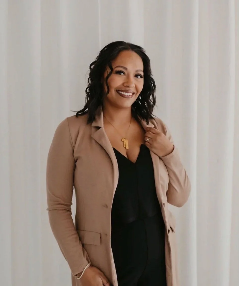 A woman with shoulder-length black curly hair, smiling, wearing a beige blazer over a black top, and a gold necklace with a number one pendant, standing against a neutral background.