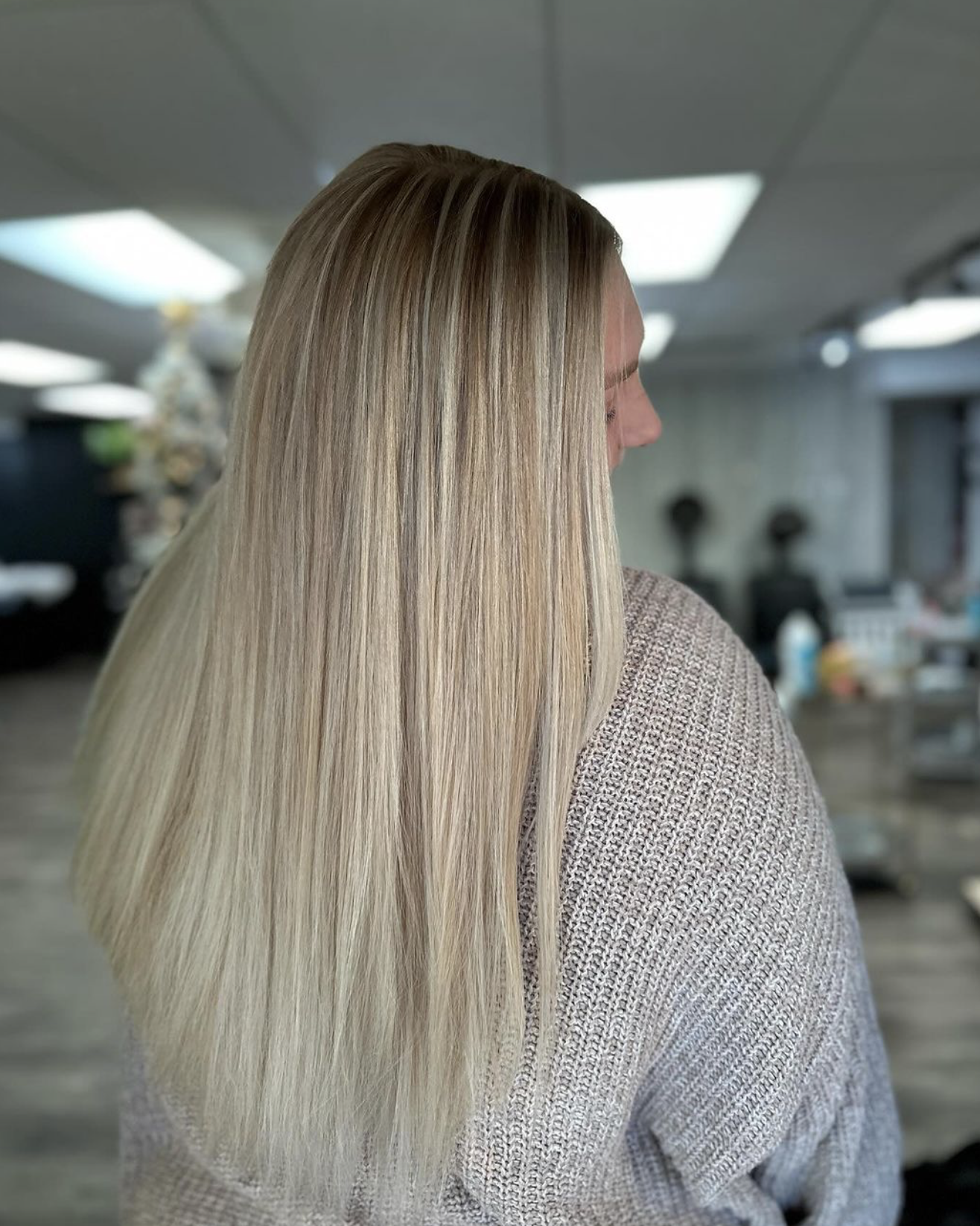 A woman with long, straight, blonde hair wearing a beige sweater in an indoor setting with ceiling lights and blurred background.