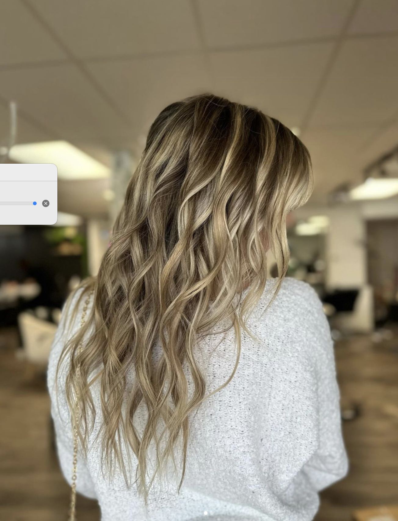 A woman with long, wavy blonde hair standing indoors with a blurred background of ceilings, lights, and furniture.