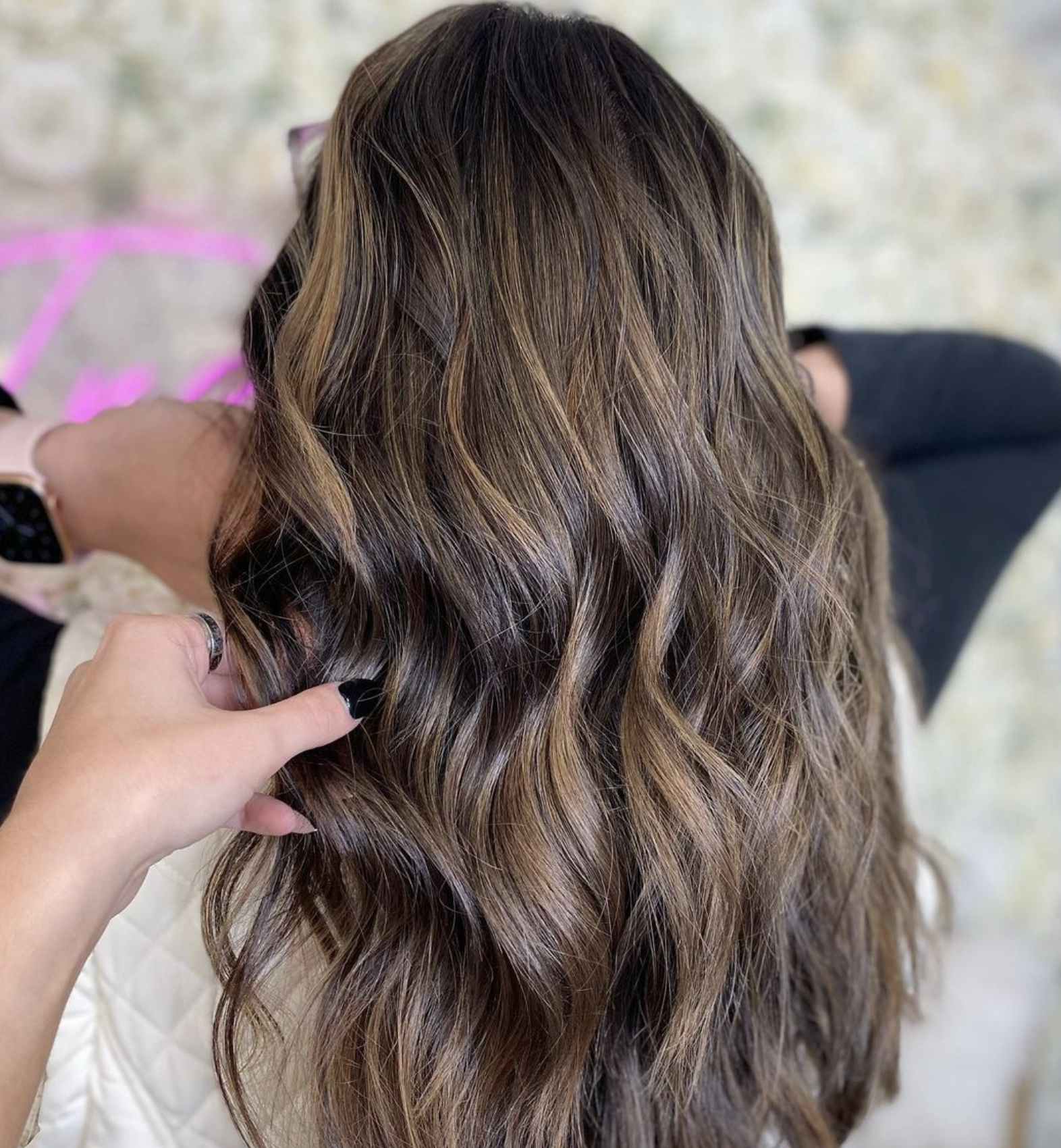 Close-up of a person with long, wavy, brown hair, held gently by someone with nail polish and rings, in a well-lit setting.