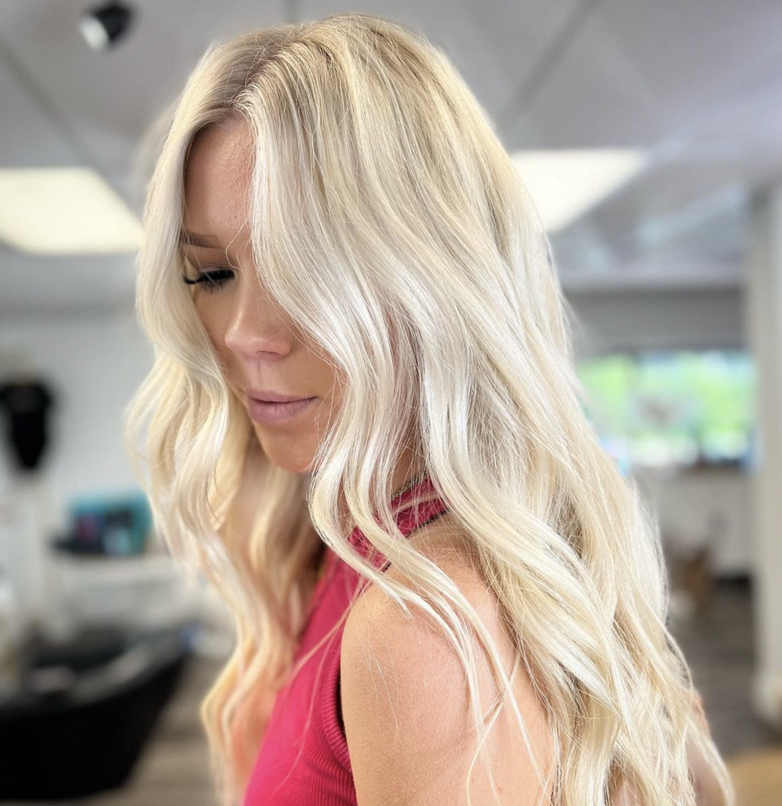 A woman with long, wavy blonde hair looking down, wearing a pink sleeveless top, in what appears to be a salon or indoor setting.