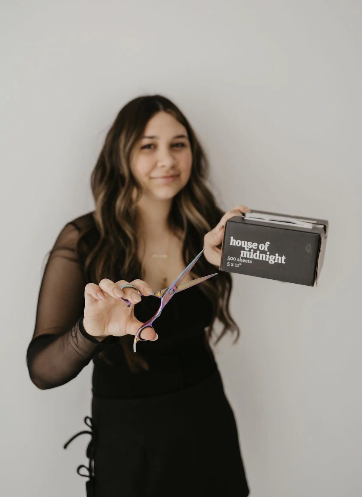 A woman holding a pair of scissors and a box of sheets labeled "house of midnight" in front of a plain white wall.