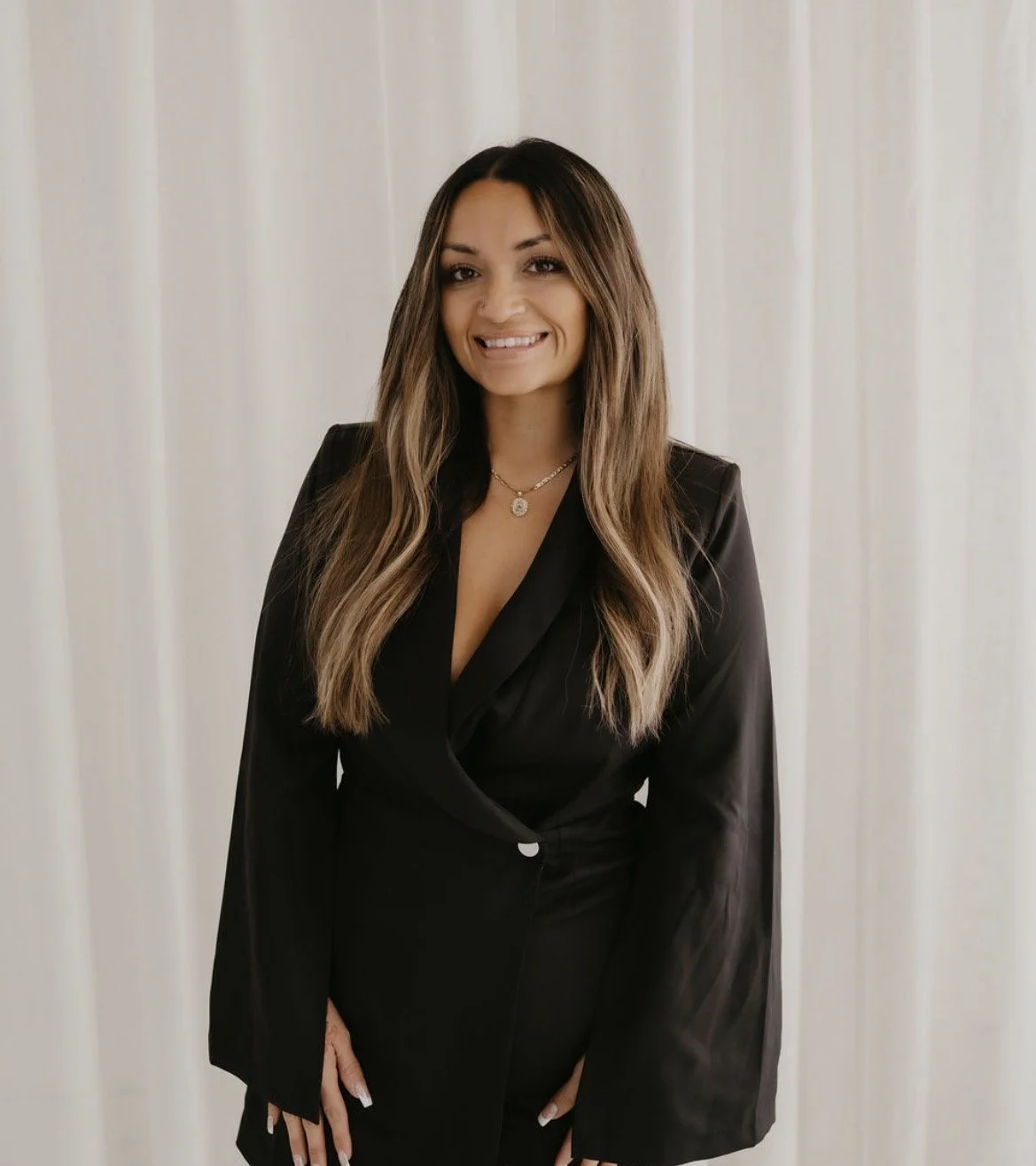 A woman with long, wavy brown hair wearing a black blazer and a gold necklace, smiling and standing in front of a plain light-colored curtain background.