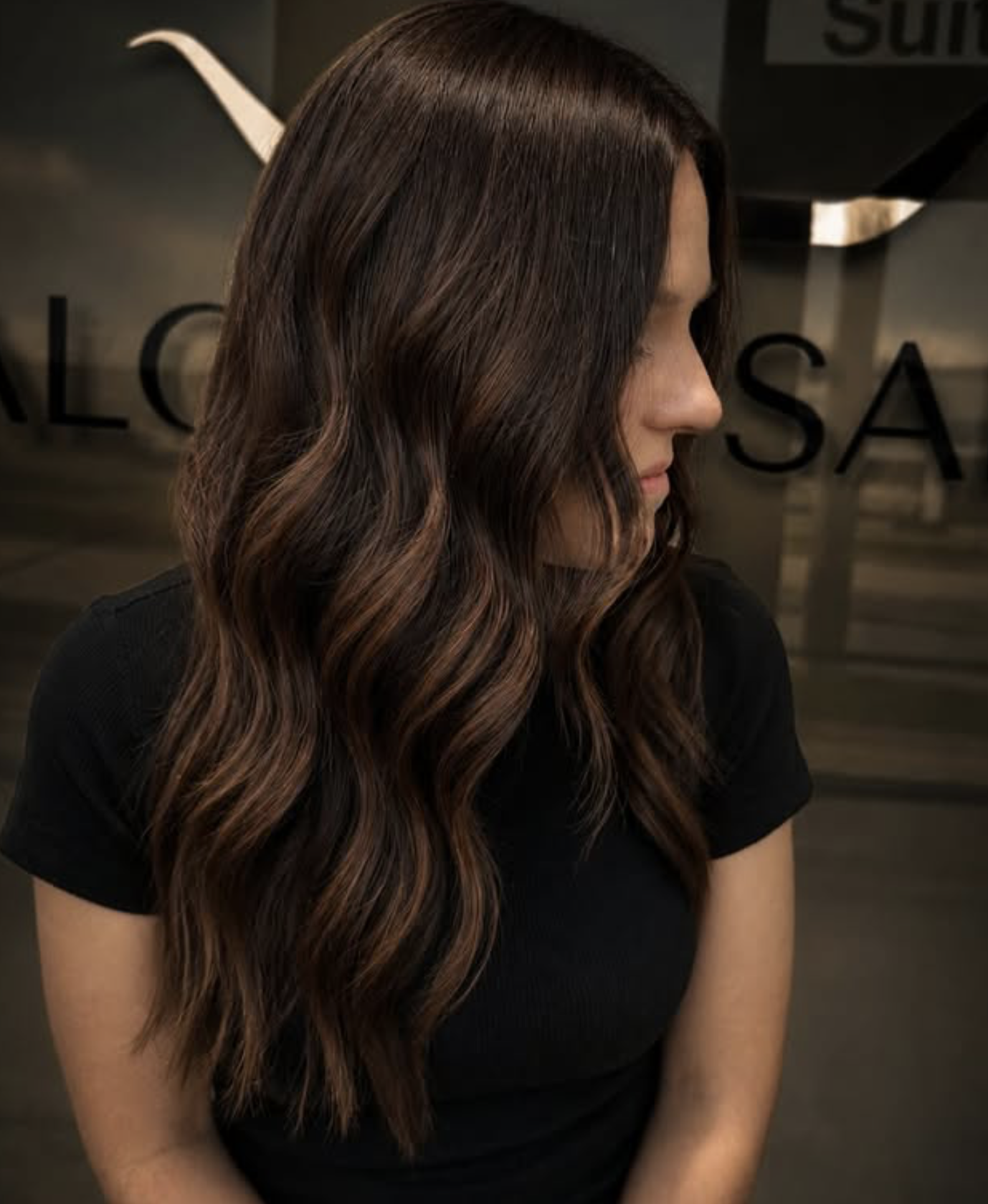 Side profile of a woman with long, wavy brown hair with lighter highlights, wearing a black shirt, standing indoors near a glass wall.