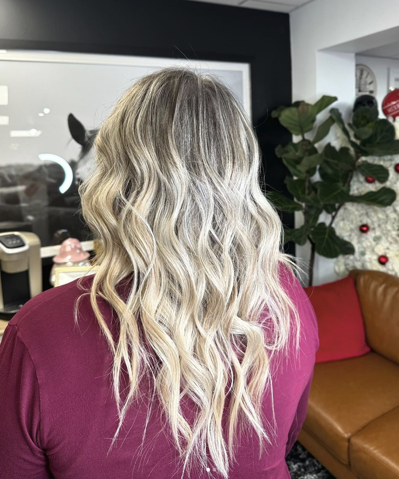 Woman with long, wavy blonde hair wearing a magenta top in a decorated indoor setting with a Christmas tree in the background.