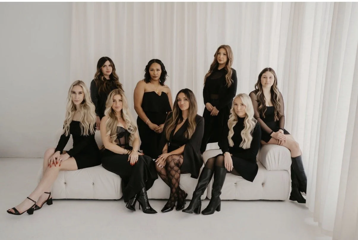 A group of nine women dressed in black outfits sitting and standing on a white sofa in front of white curtains.