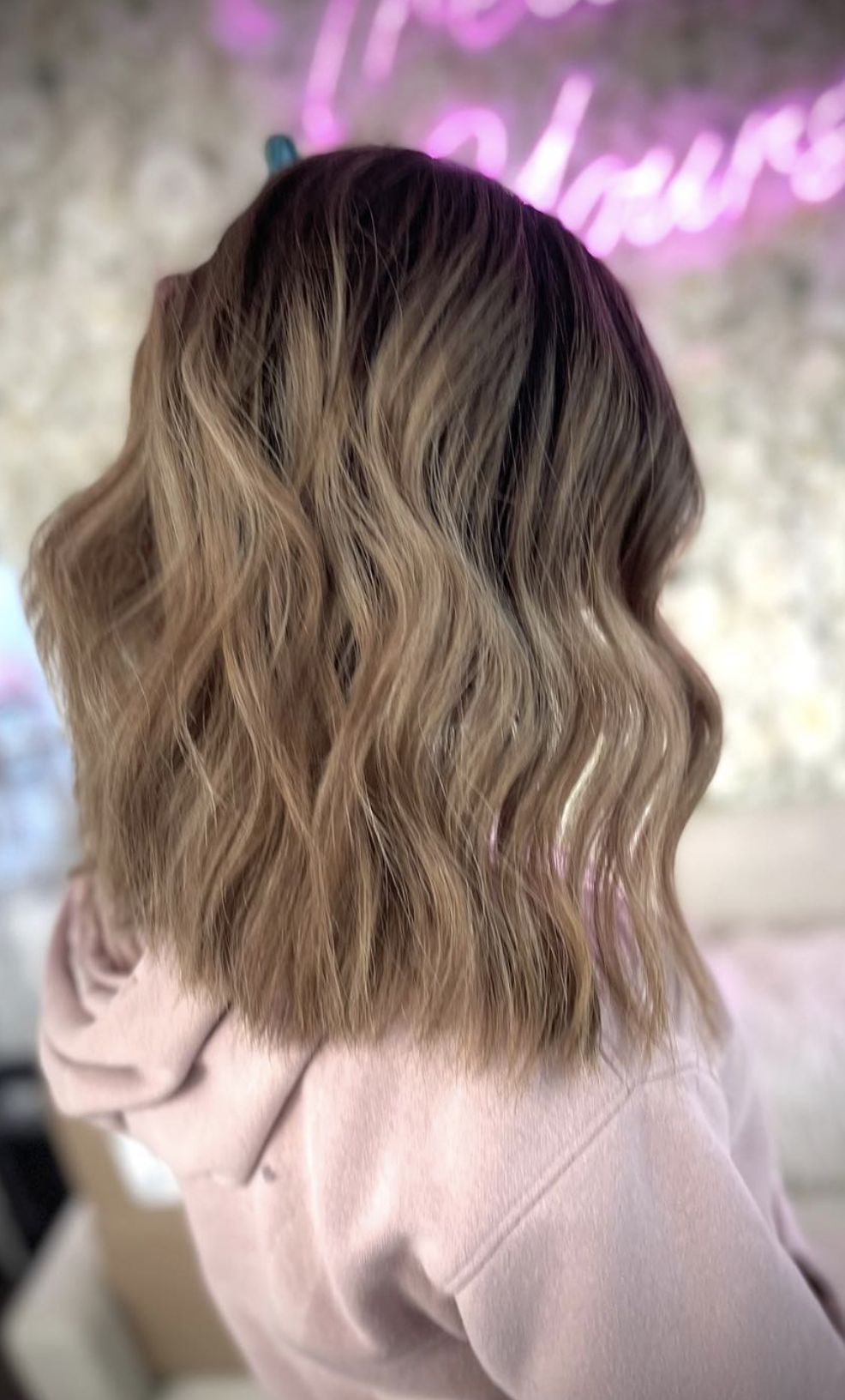 The image shows a person with shoulder-length wavy hair with blonde highlights, wearing a light pink hoodie.