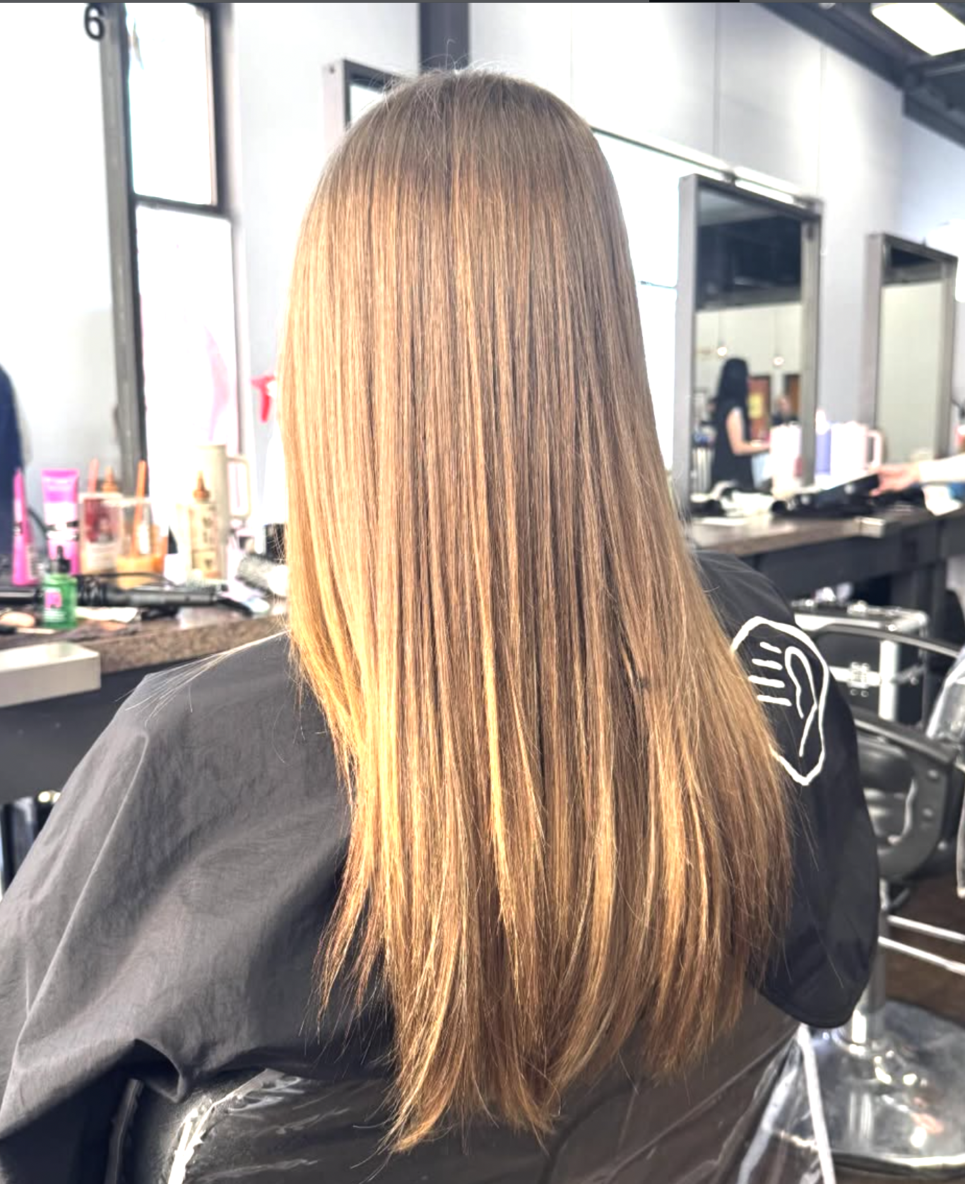 A person with long, straight, honey-colored hair sitting in a salon chair, getting a hair treatment or haircut, with salon equipment and mirrors visible in the background.