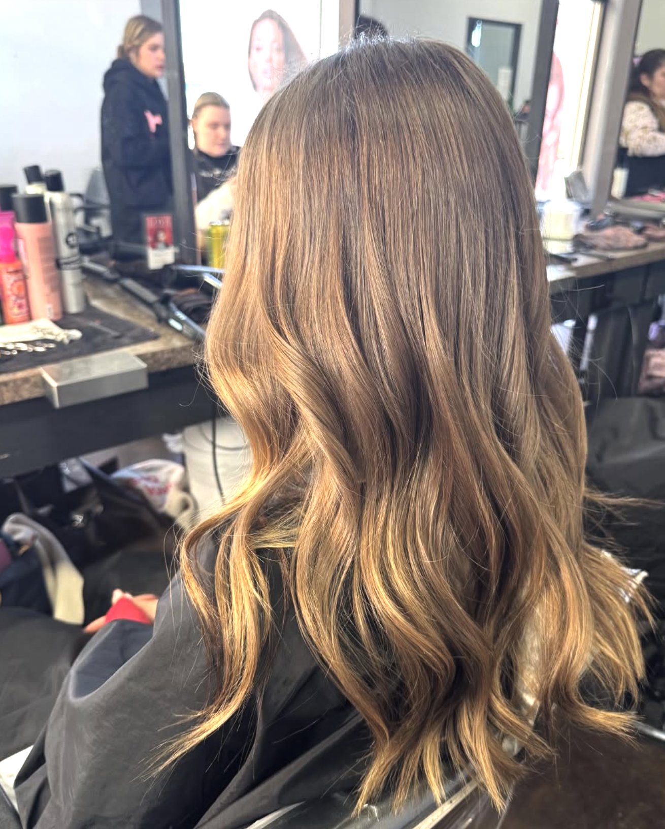 A woman with long, wavy, caramel-colored hair seated at a salon, with mirror and hair styling products visible in the background.
