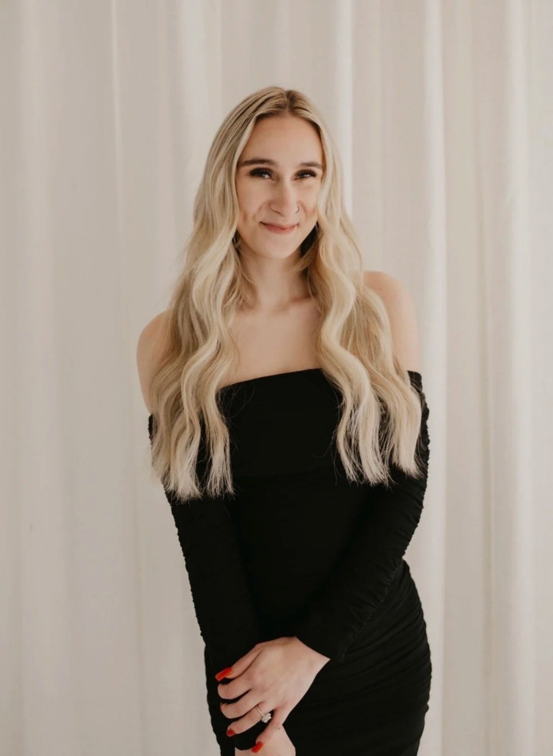 A woman with long wavy blonde hair wearing a black off-the-shoulder dress, standing in front of white curtains, smiling softly.