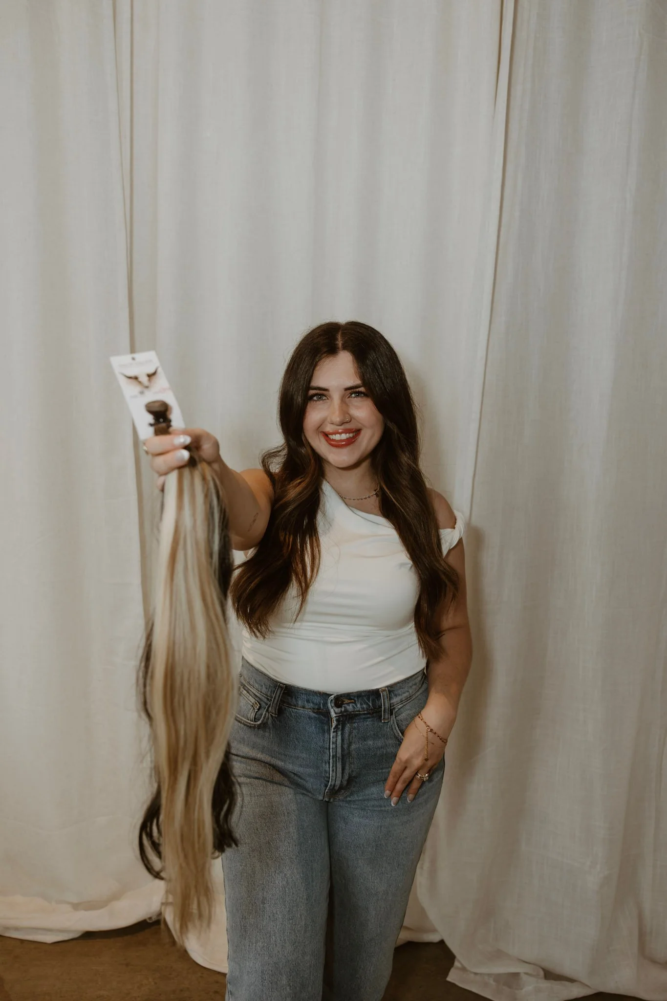 A woman with long, wavy brown hair smiling and winking, wearing a black top, black pants, and a black blazer, standing in front of white curtains.