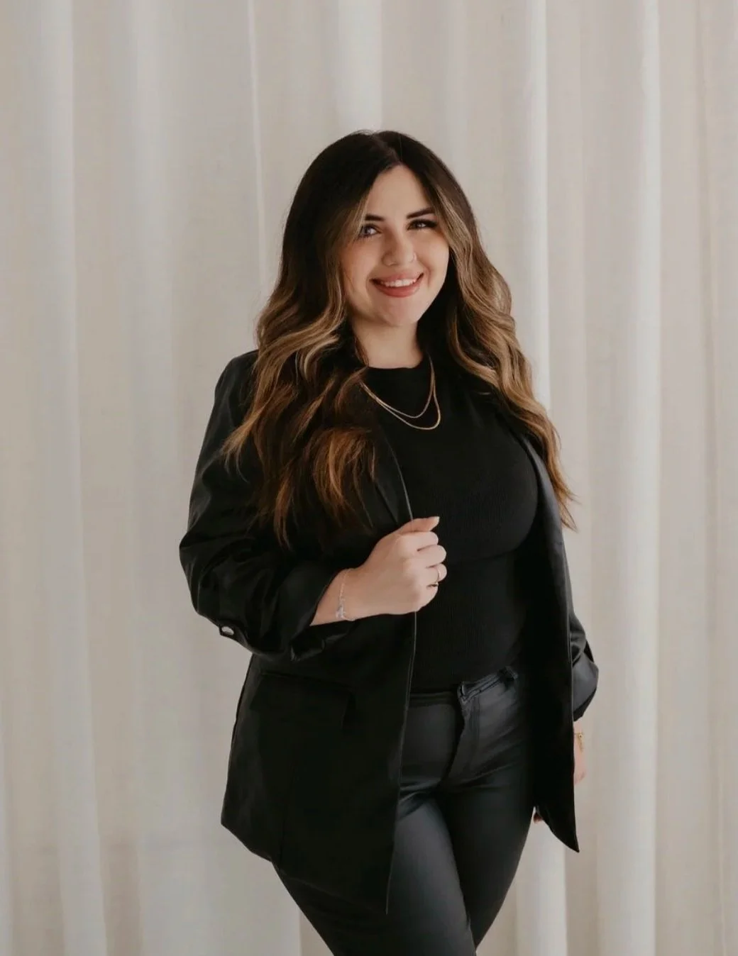 A woman with long, wavy brown hair smiling and winking, wearing a black top, black pants, and a black blazer, standing in front of white curtains.