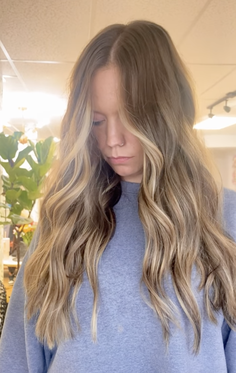A young woman with long, wavy blonde hair with darker roots, looking downward, wearing a gray sweatshirt, indoors with warm lighting and a green plant in the background.