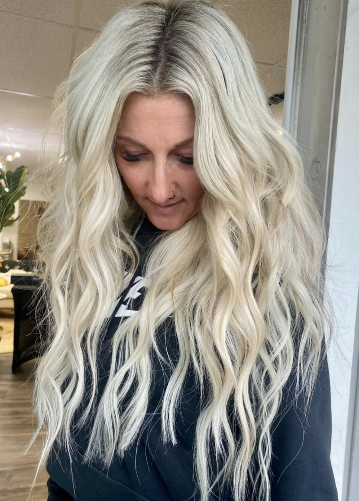 A woman with long, wavy platinum blonde hair and a nose piercing looking down in an indoor setting.