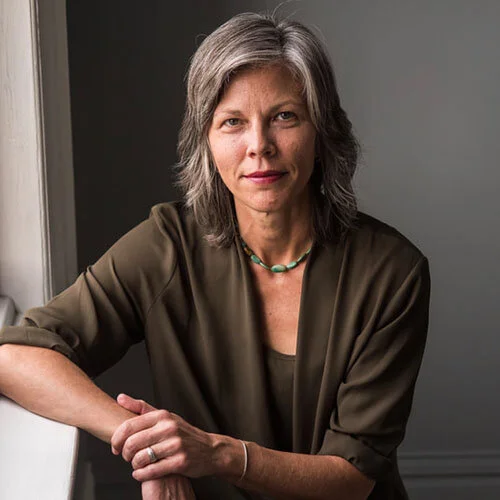 A middle-aged woman with gray hair, wearing a brown blouse and a green necklace, sitting with her arm resting on a surface near a window.