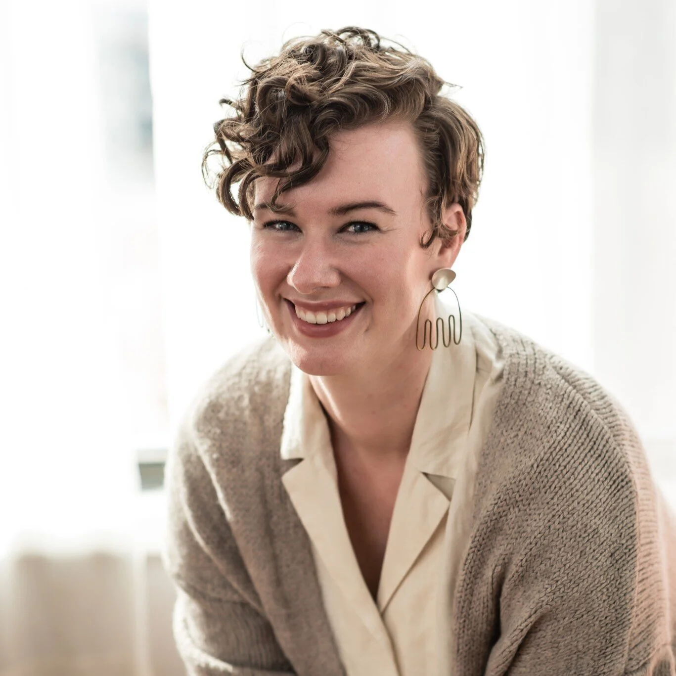 A woman with short curly hair smiling, wearing large gold earrings and a beige cardigan over a light-colored blouse, in a well-lit indoor setting.
