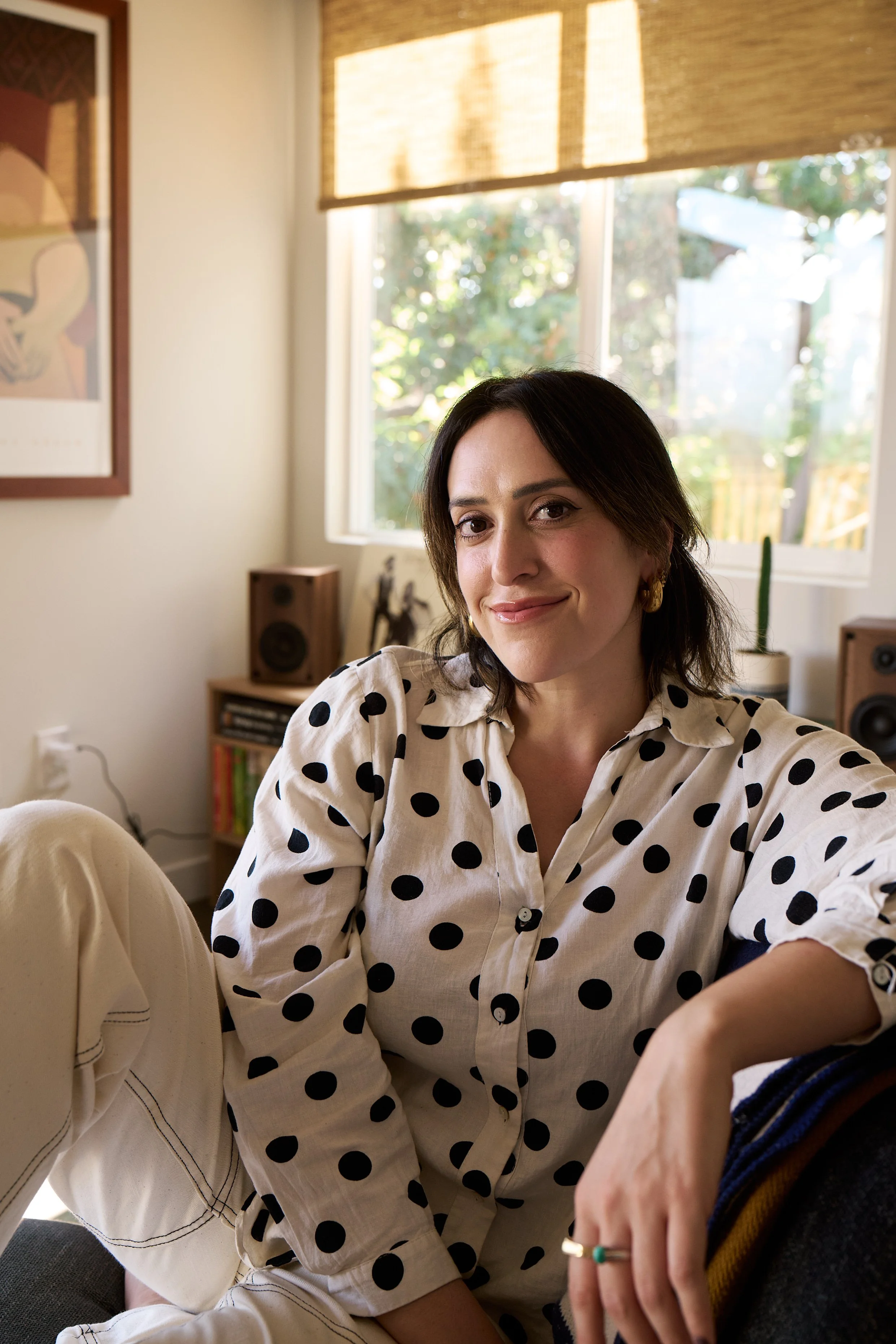 A woman with dark hair wearing a white shirt with black polka dots, sitting indoors with sunlight coming through a window behind her.