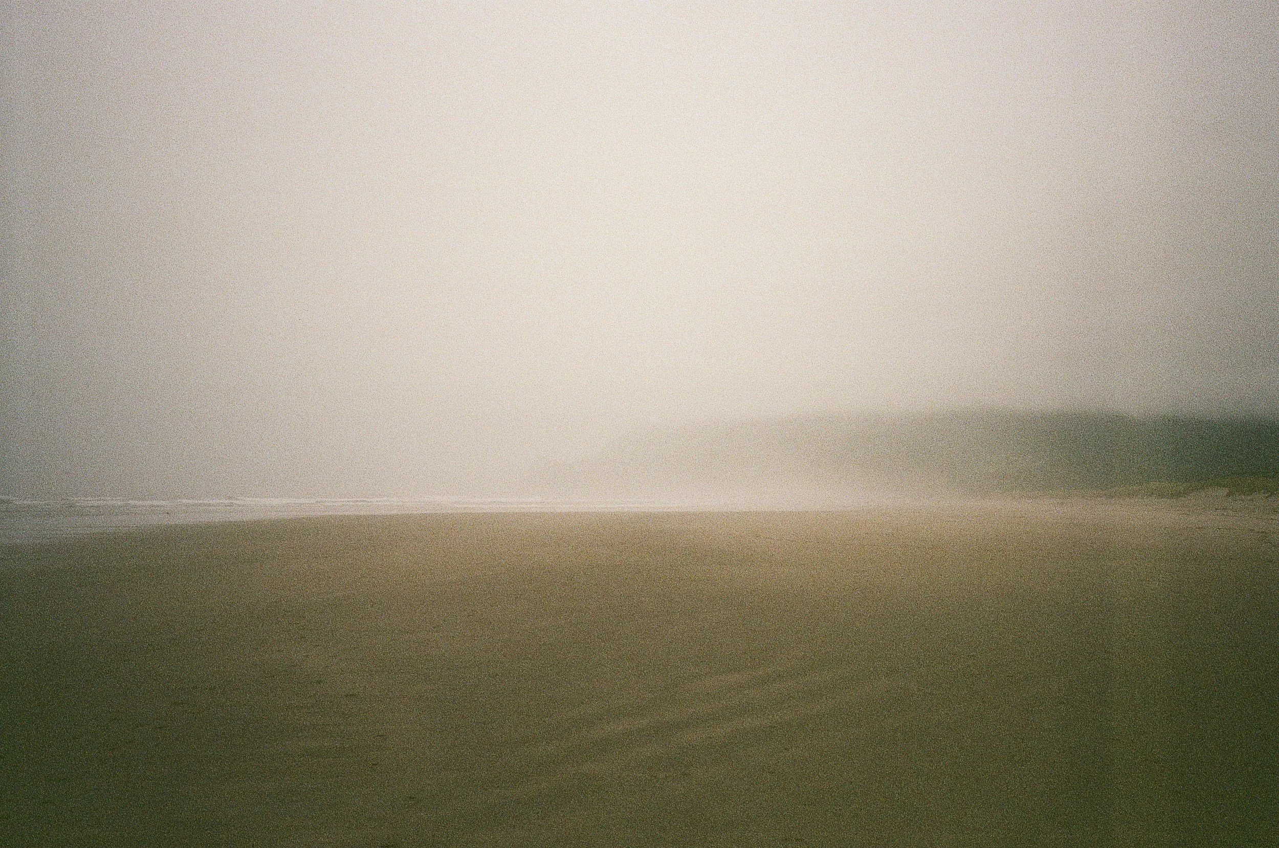 A foggy beach with sand in the foreground and the ocean in the background, partially obscured by thick fog.