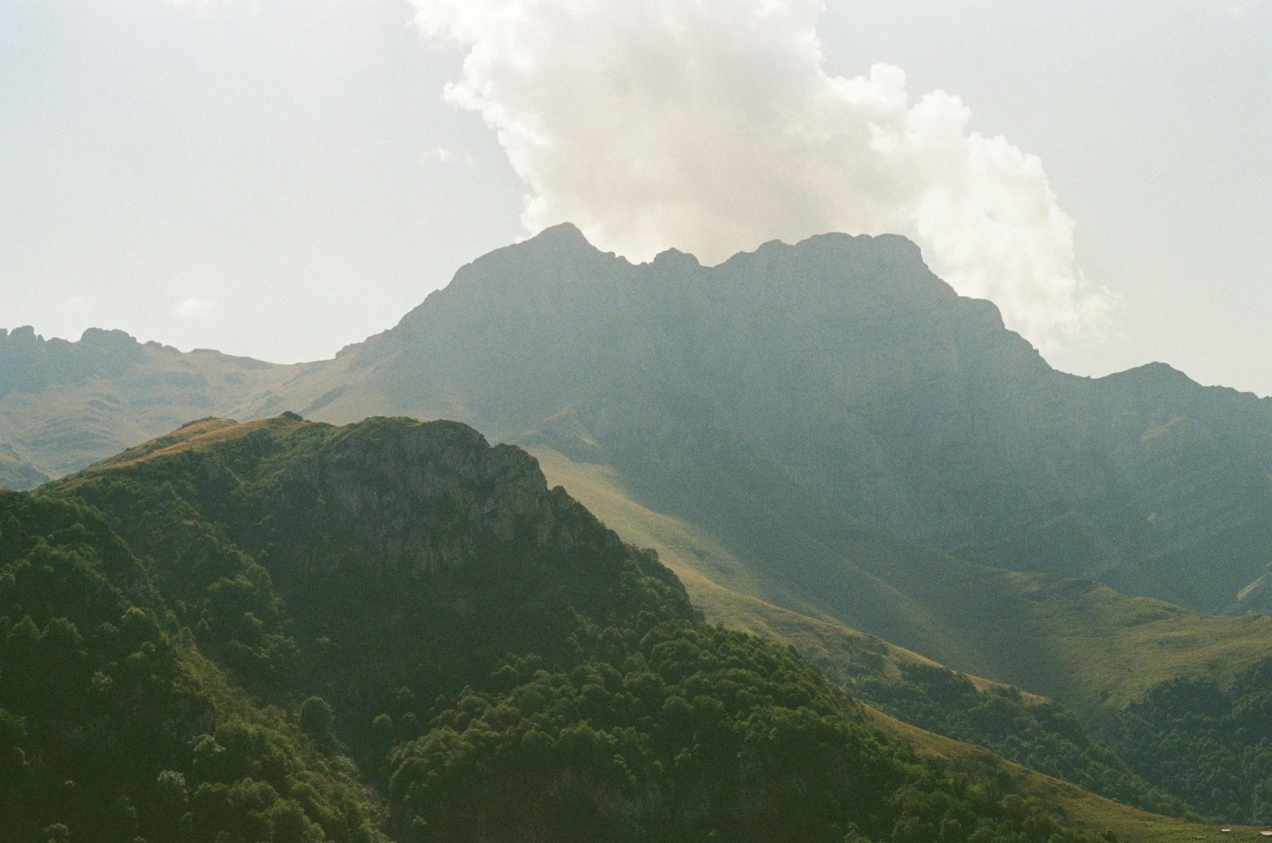 Mountain landscape with lush green hills in the foreground, rocky mountain ridges in the middle, and a mountain with clouds behind.