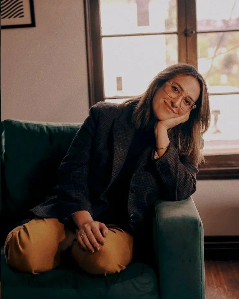 A woman with glasses and shoulder-length hair sitting on a green couch, resting her head on her hand, in a room with a window and natural light.