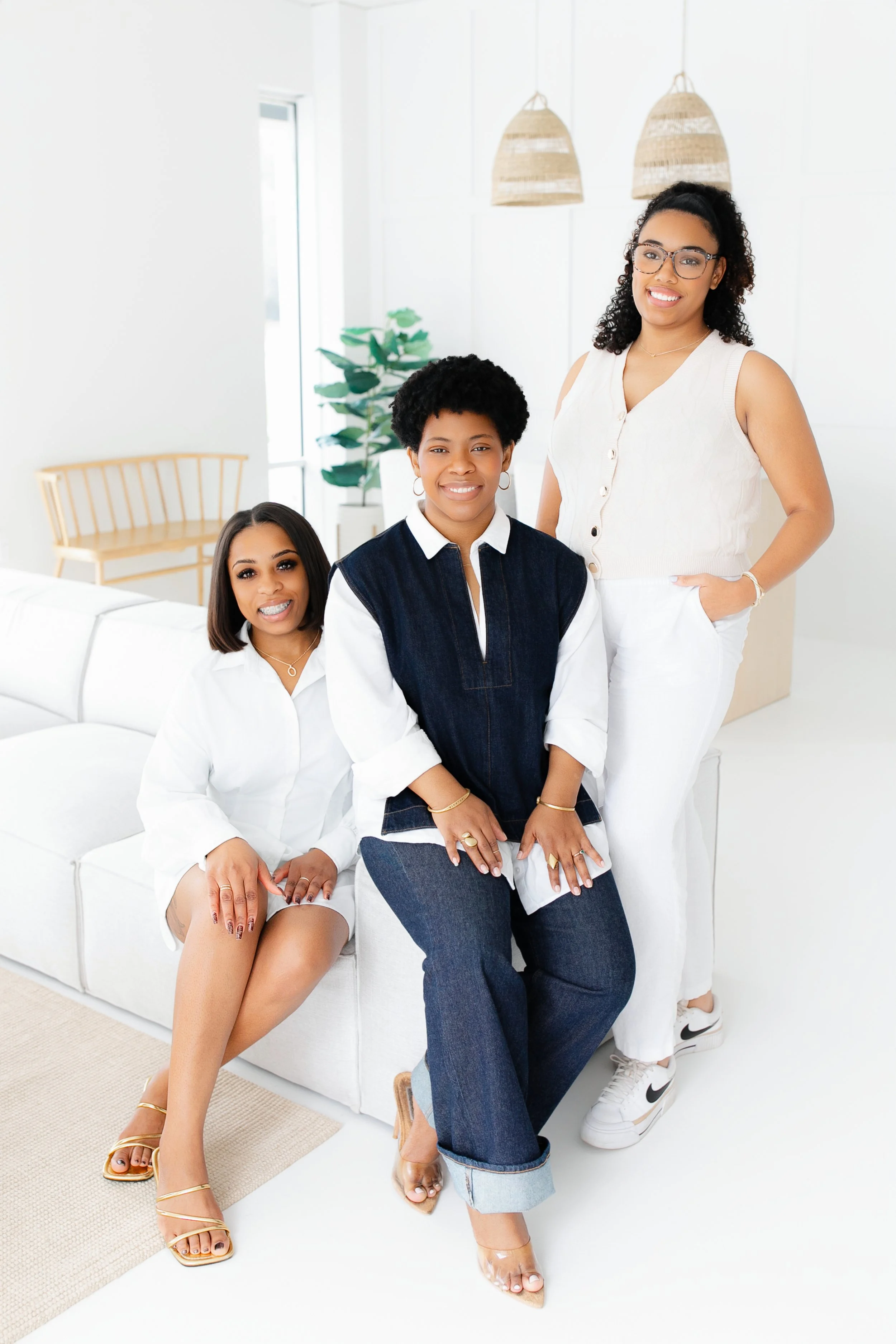 Three black African American women in a bright white room, smiling and posing for a team photoshoot. The room has minimal decor, including a plant and hanging lamps. Female therapists of Jacksonville, FL - Duval County