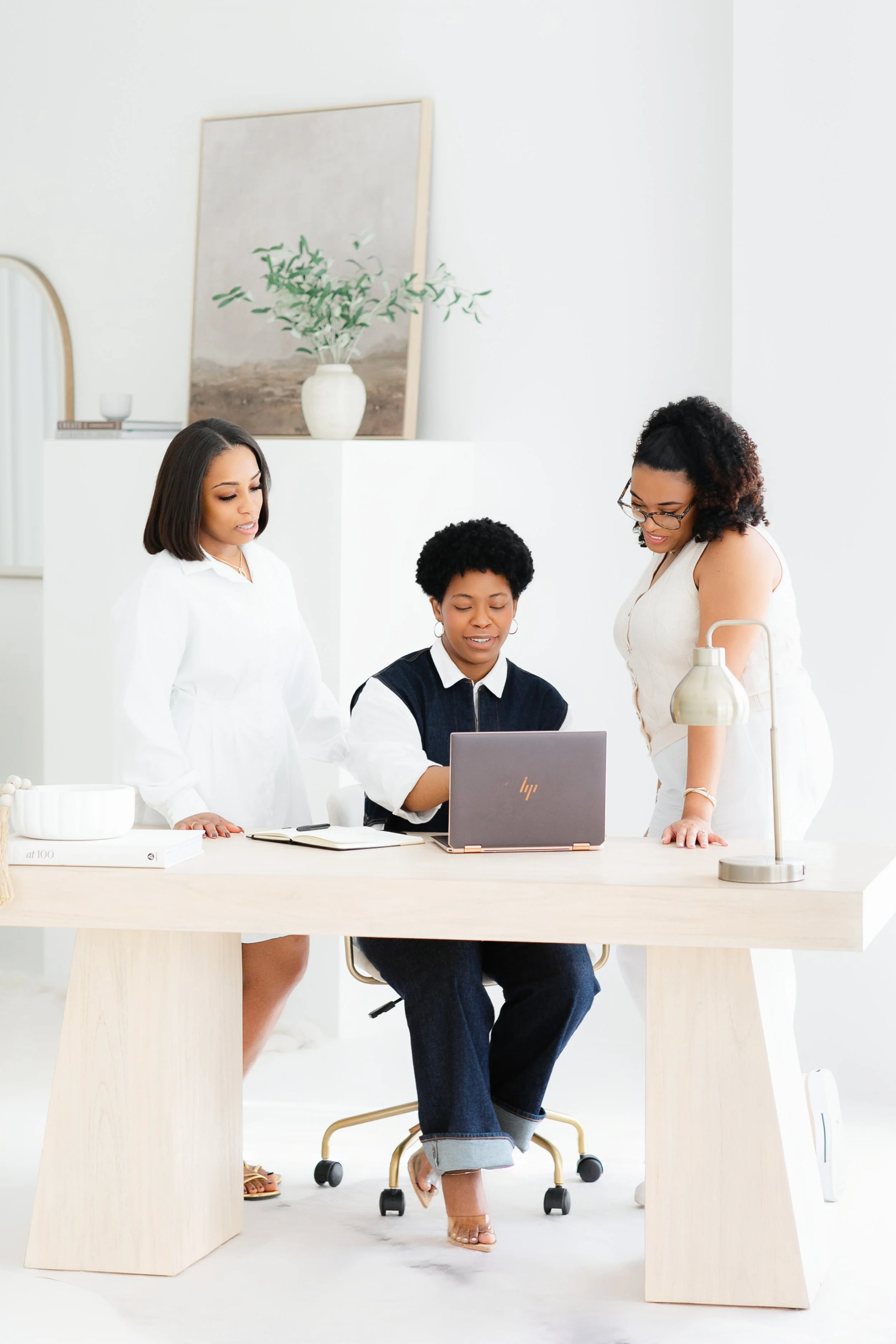 Three Black women in a bright, modern office space, gathered around a laptop on a light-colored desk, with a white background and decorative plant and artwork.  Professional photoshoot idea for Pinterest.  Female therapists of Jacksonville, FL