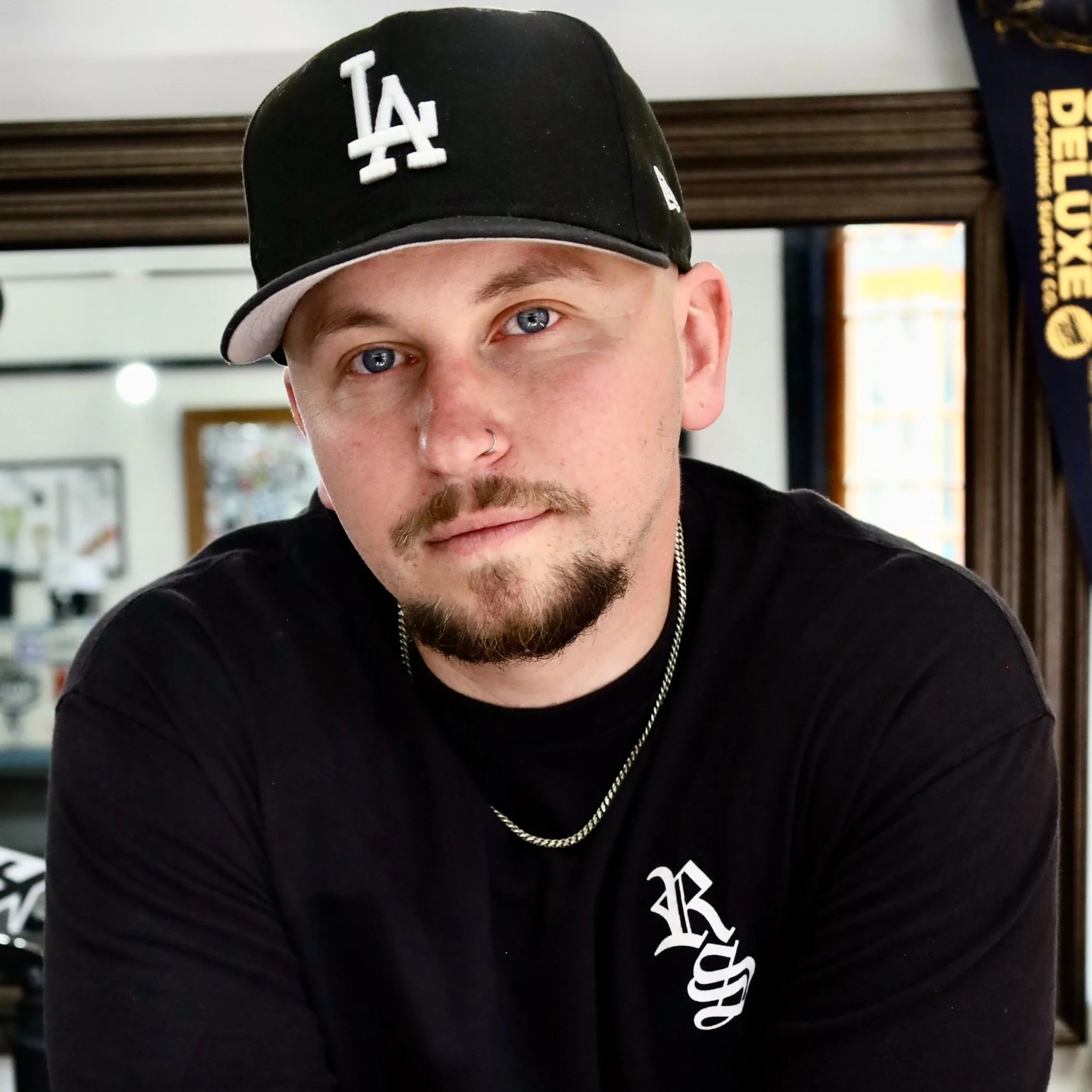 Shop owner and barber, Austin Jacquemin, wearing a black LA Dodgers cap, black T-shirt with white lettering, a silver chain, and a nose piercing, looking at the camera indoors.