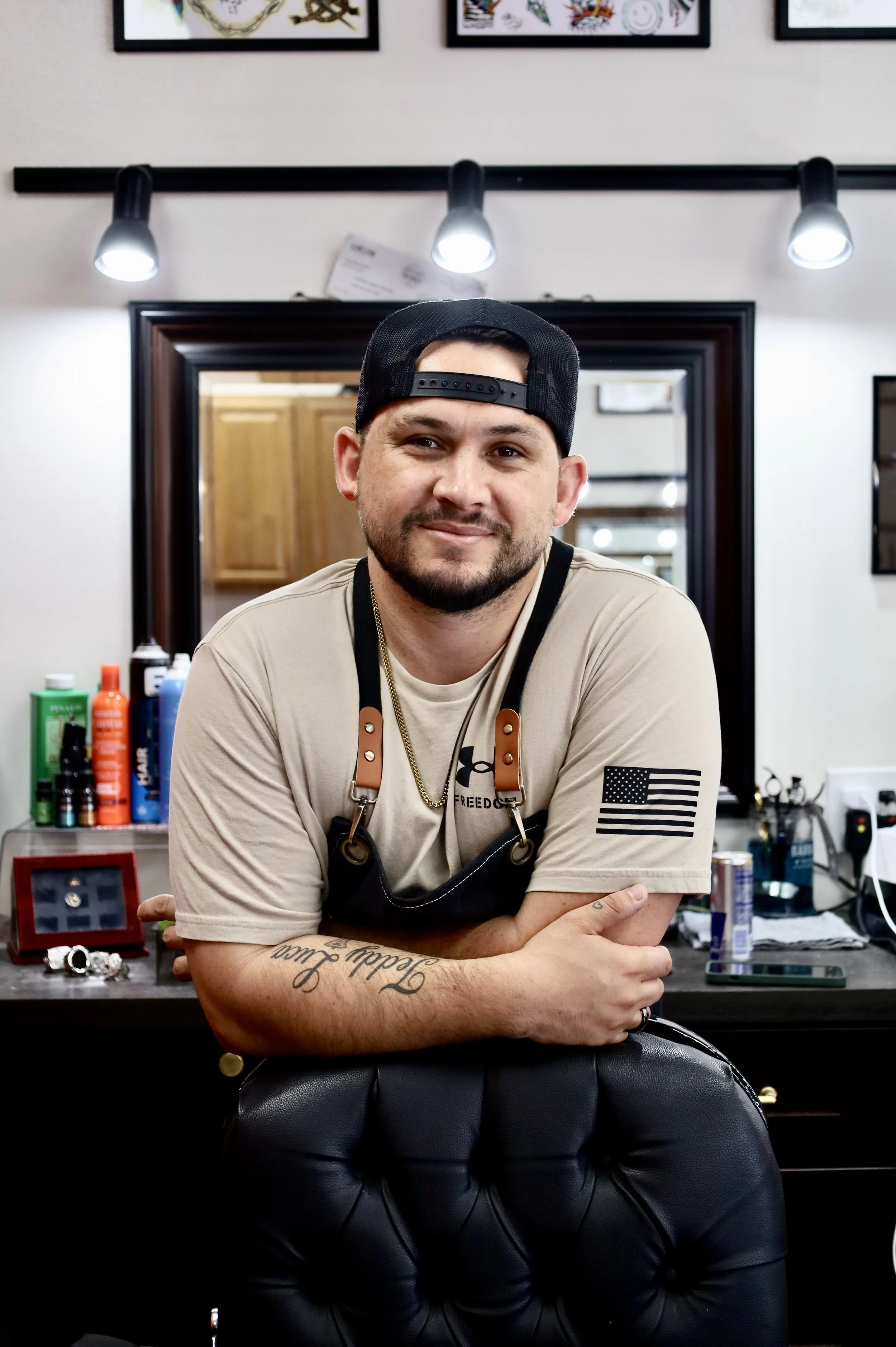 A man with a beard wearing a black backward cap, a beige t-shirt with an American flag patch on the sleeve, and a gold chain, leaning on a black barber chair inside a barbershop with grooming products in the background.