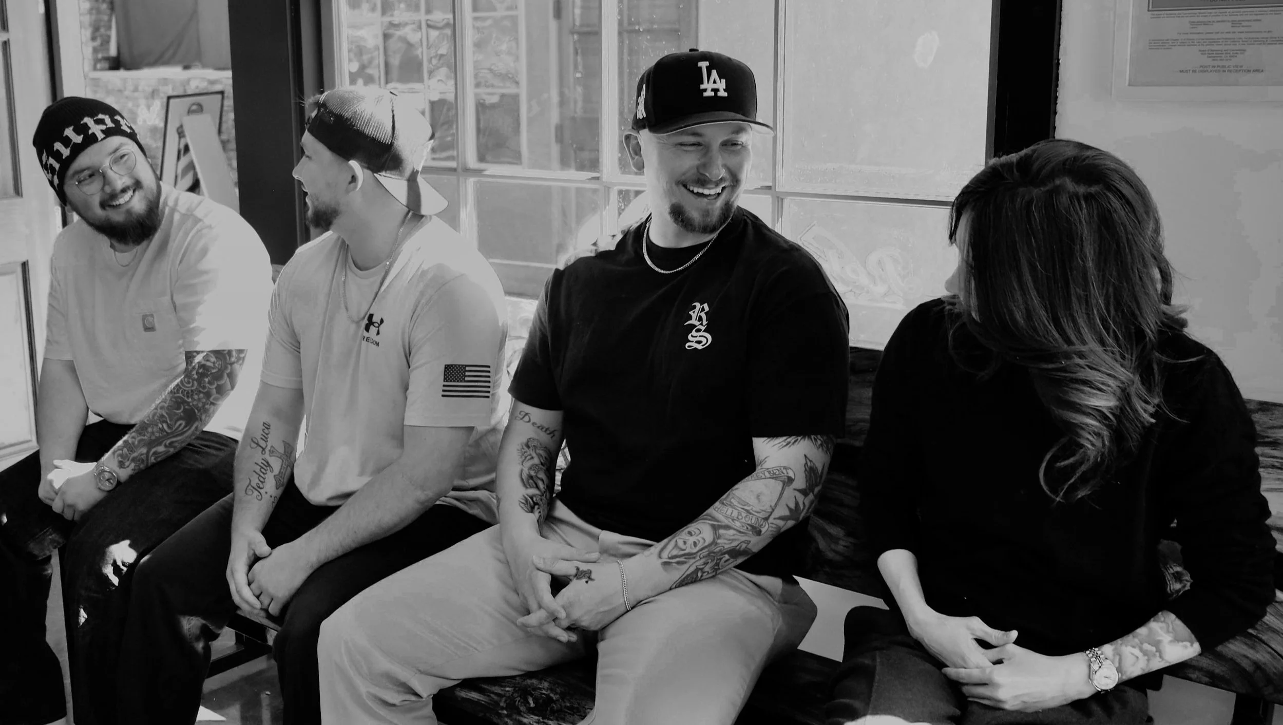 Four people sitting on a bench at a cafe, smiling and conversing. The group consists of three men and one woman. Two of the men are wearing caps, and all have tattoos on their arms. They are engaged in a lively conversation, with natural light streaming in from behind the window.